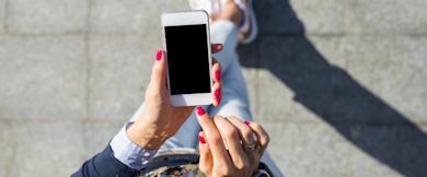 A person with pink nail polish holds a white smartphone with a blank screen, using it outdoors on a paved surface, wearing light blue jeans and white shoes.
