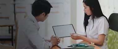 A female doctor shows a blank tablet screen to a male patient during a consultation at a desk with a stethoscope and documents.