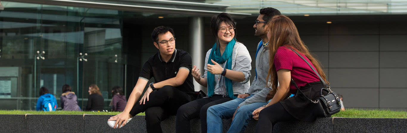 Diverse group of students sitting and chatting on a concrete ledge outside a modern glass building on campus