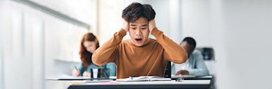 A person sitting in classroom, at a desk, with their hands to their head looking overwhelmed