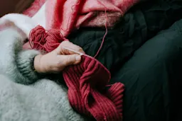 En hånd strikker med rødt garn, omgitt av myke, fargerike pledd.