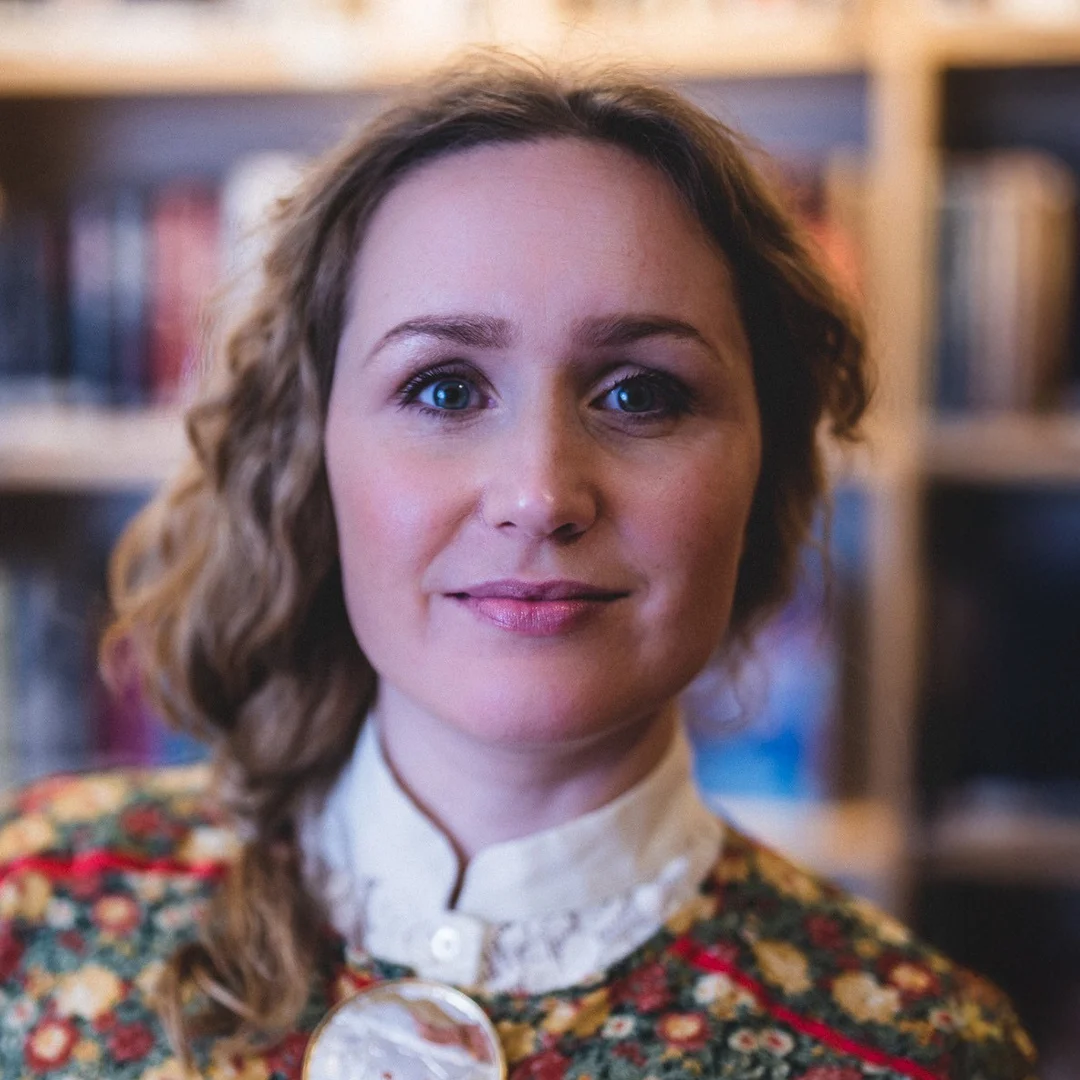 Forfatterportrett av Tina Harnesk i blomsterkjole med smykker foran en bokhylle.