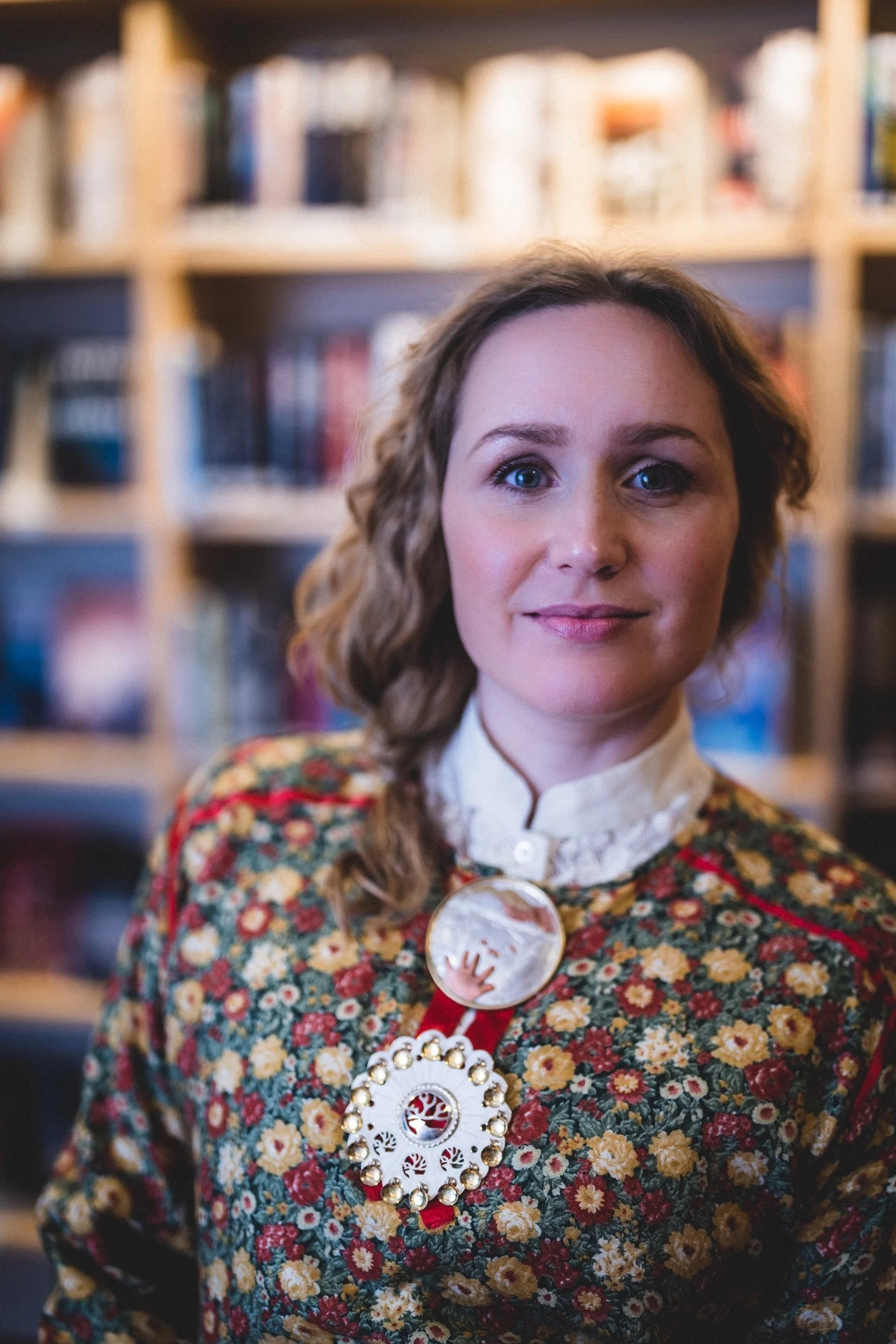 Forfatterportrett av Tina Harnesk i blomsterkjole med smykker foran en bokhylle.