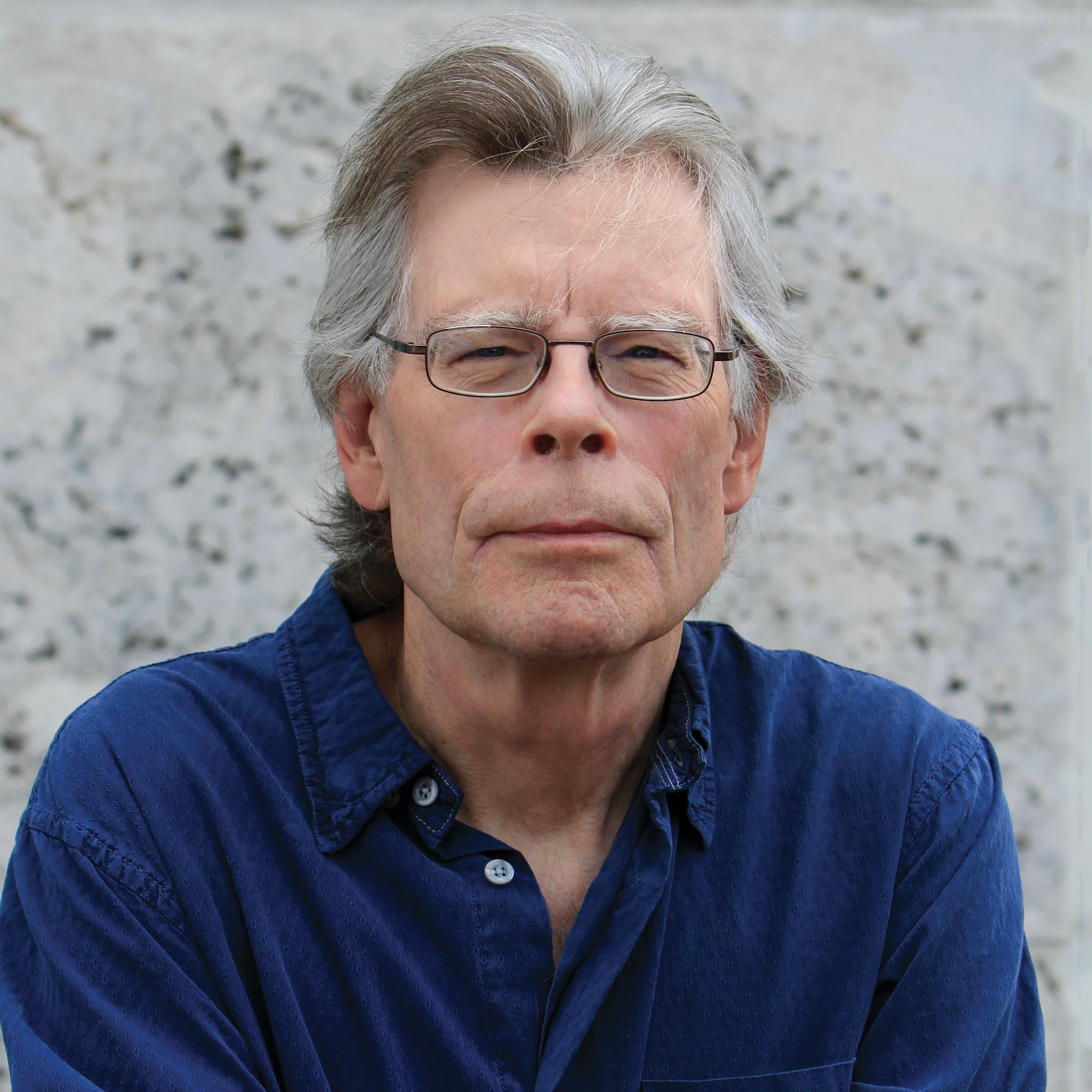 Porträtt av författaren Stephen King, klädd i en blå skjorta, sittande mot en ljus bakgrund.