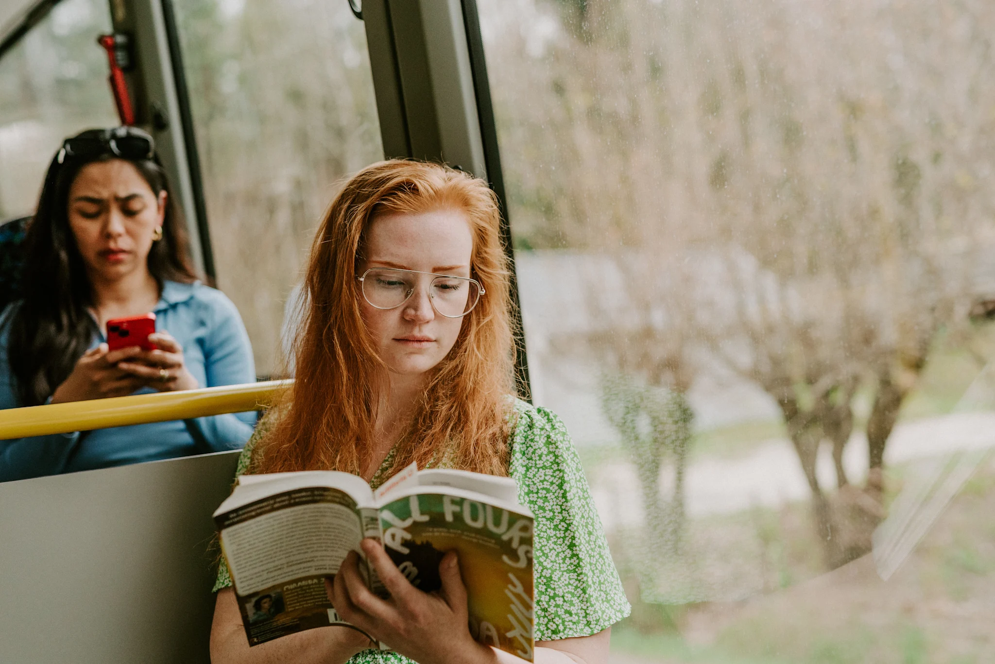 En person med rødt hår og grønn kjole sitter på en buss og leser en bok.