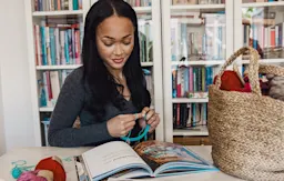 En person som stickar med en bok om stickning och en korg med garn i bakgrunden.