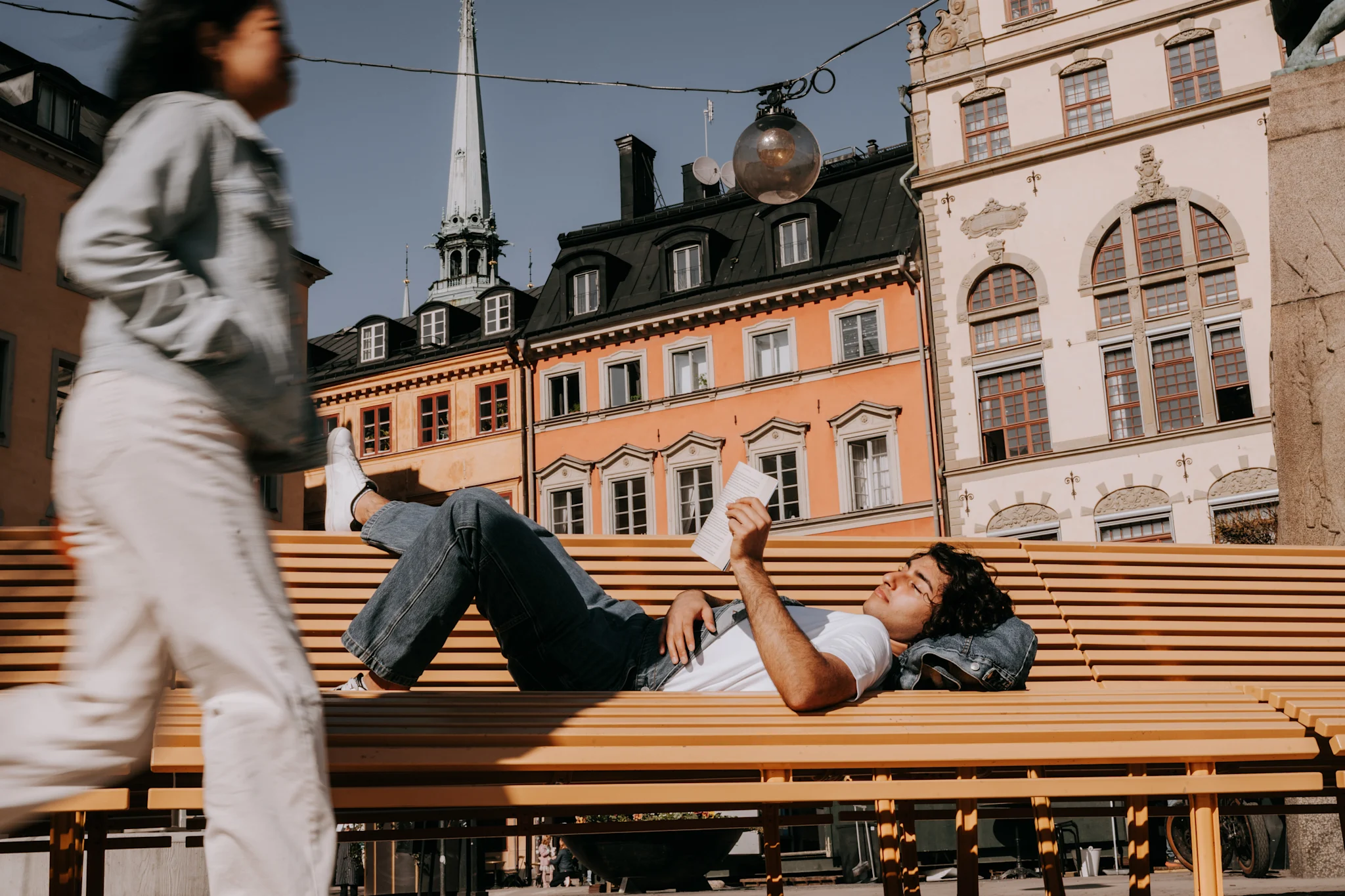 En person ligger på en bänk och läser en bok, omgiven av byggnader i en stadsmiljö.