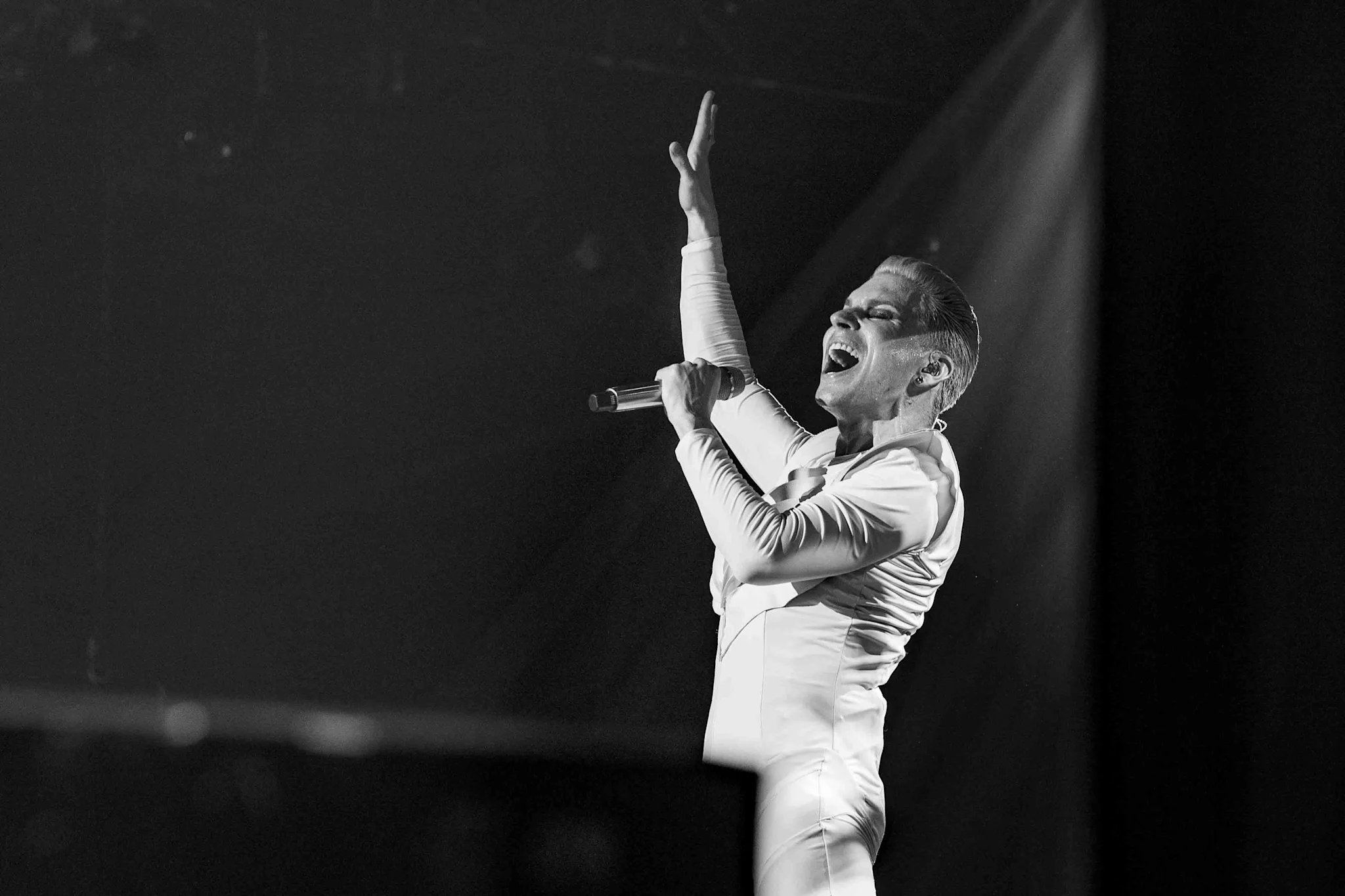Esityksessä oleva artisti valkoisissa vaatteissa mikrofoni kädessään, nostaa kätensä konserttitilanteessa.