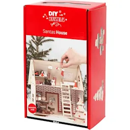 DIY-sett for å bygge et juletrehus, med en hånd som plasserer en dekorasjon.