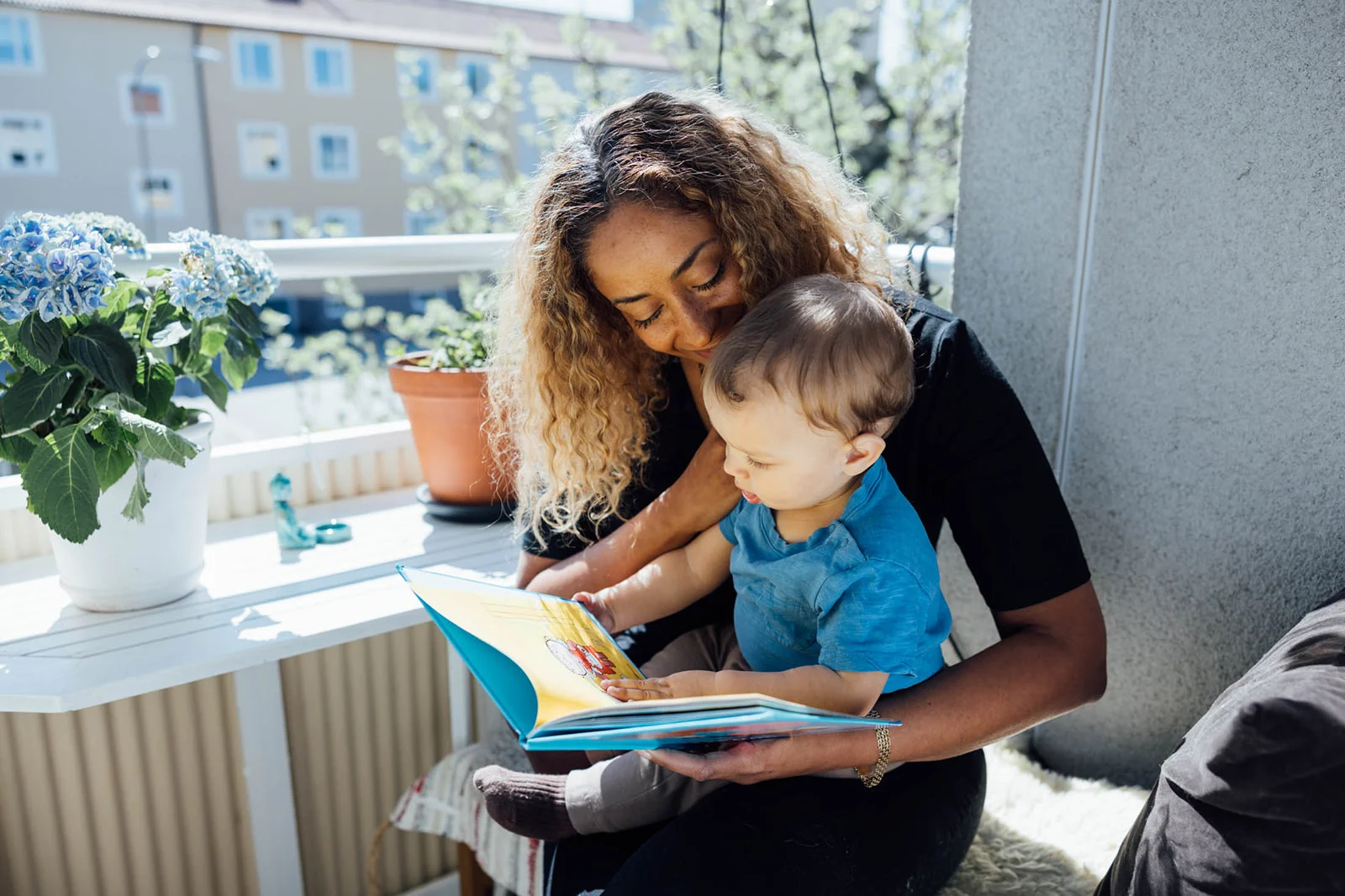 En vuxen och ett barn läser en bok tillsammans på en balkong med blommor i bakgrunden.