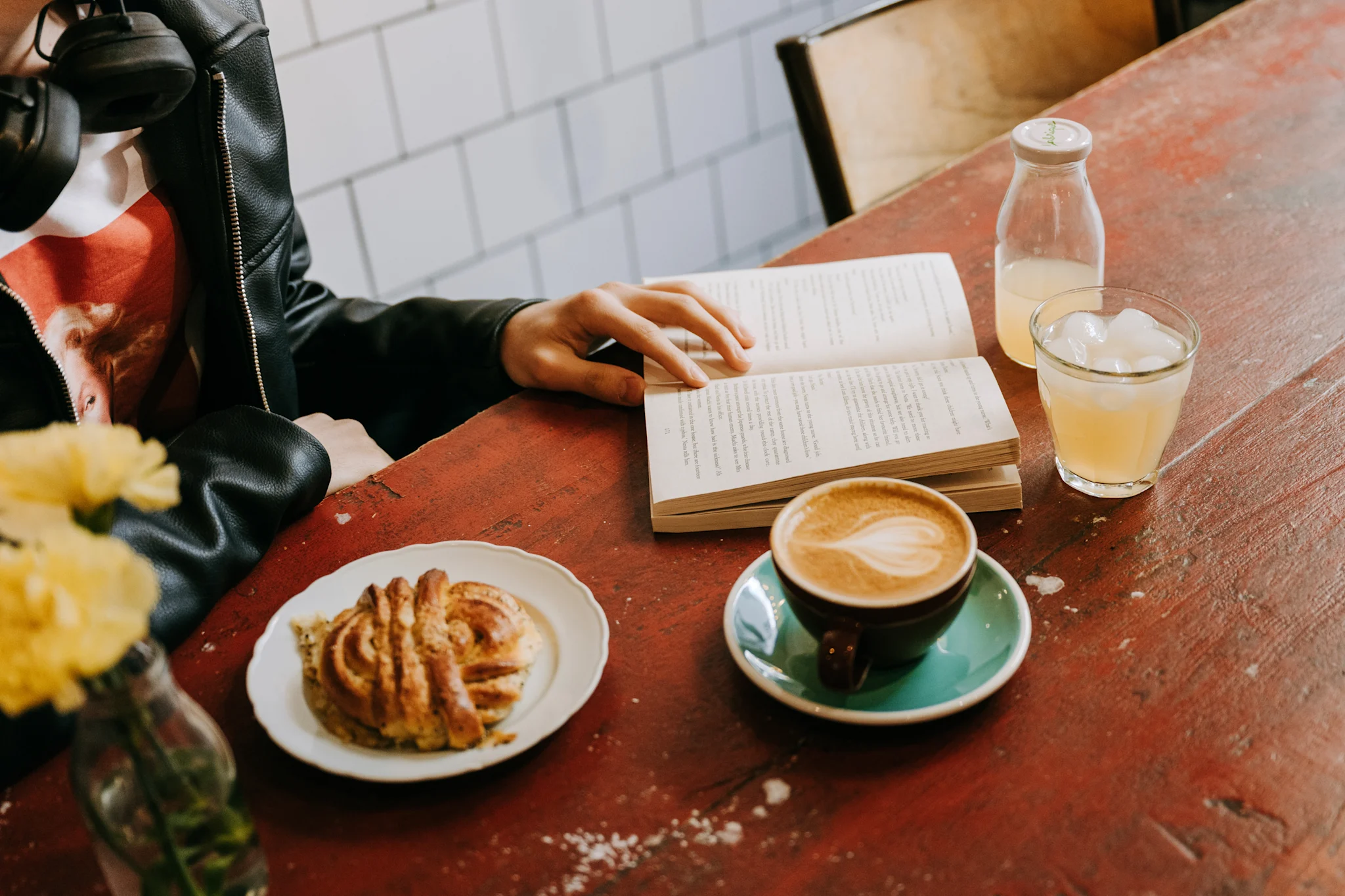 En person sitter ved et bord med en bok, kaffe, en drink og en bolle på en rød treoverflate.