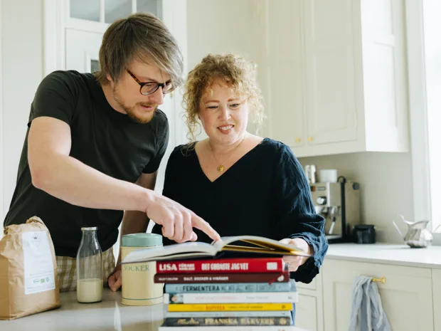 To personer i et kjøkken diskuterer matlaging med ferske grønnsaker og en kokebok på bordet.