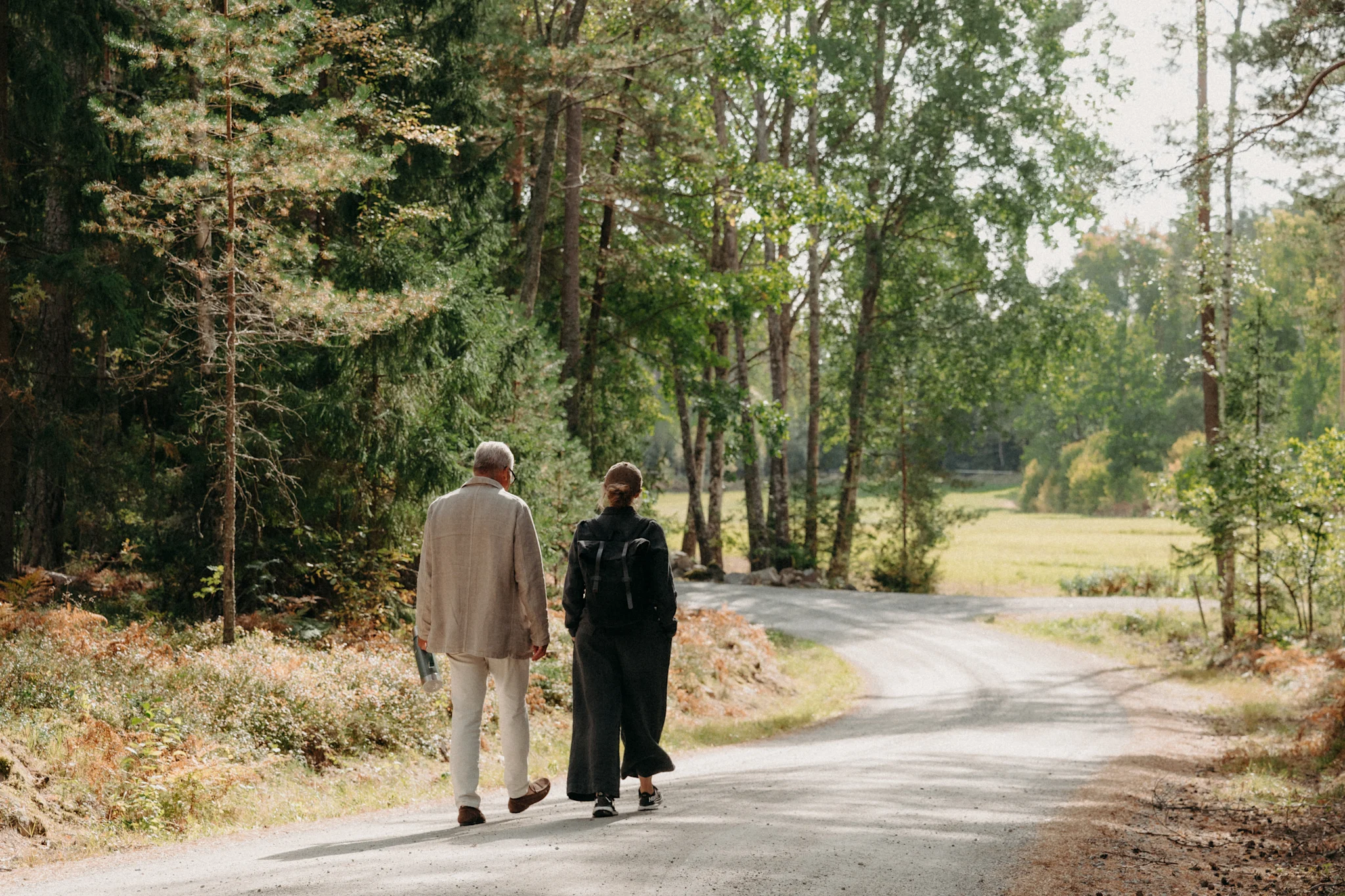 Kaksi henkilöä kävelee soratiellä, jota ympäröivät puut ja viheralueet.