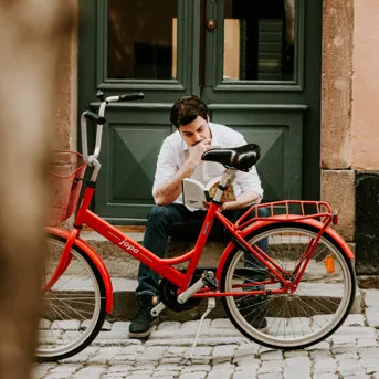 En person sitter på en gata och läser en bok bredvid en röd cykel.