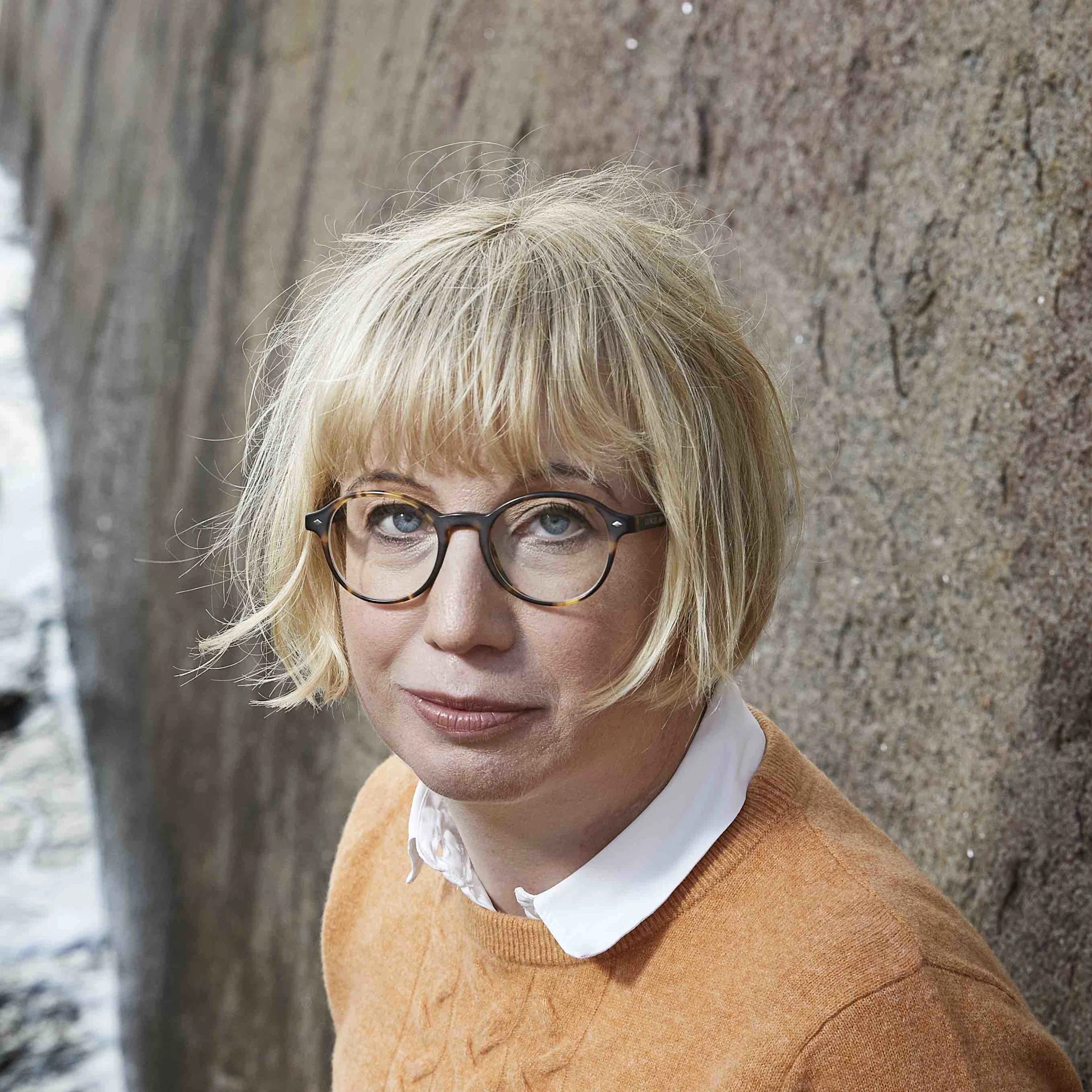 Kirjailija Kristina Ohlssonin muotokuva, istuu seinää vasten, vaaleat hiukset ja oranssi neule.