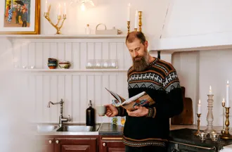 En man med skägg står i ett kök och läser i en bok medan han håller en kopp.