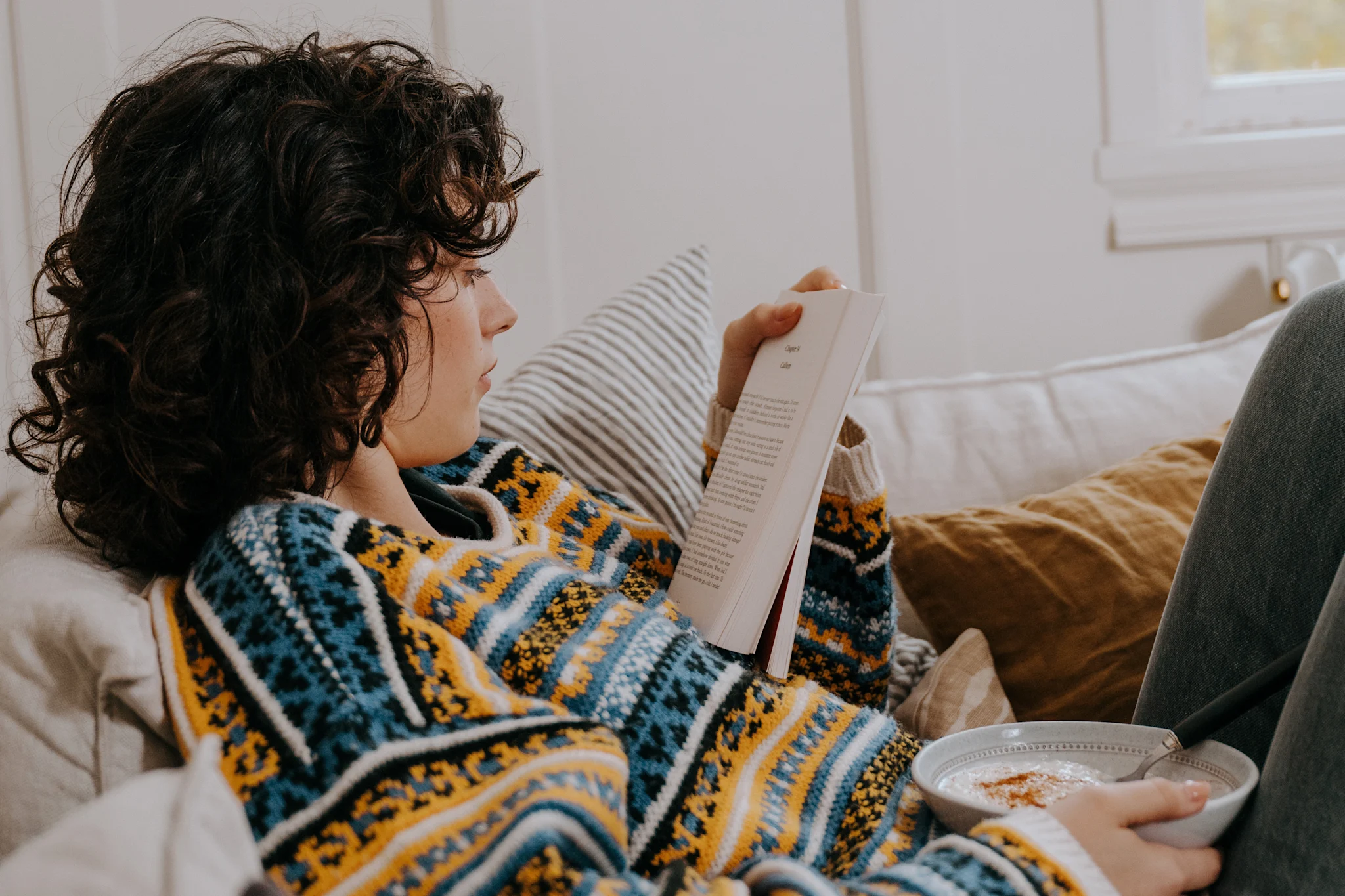 En person sitter i en soffa och läser en bok medan de håller en skål med mat.