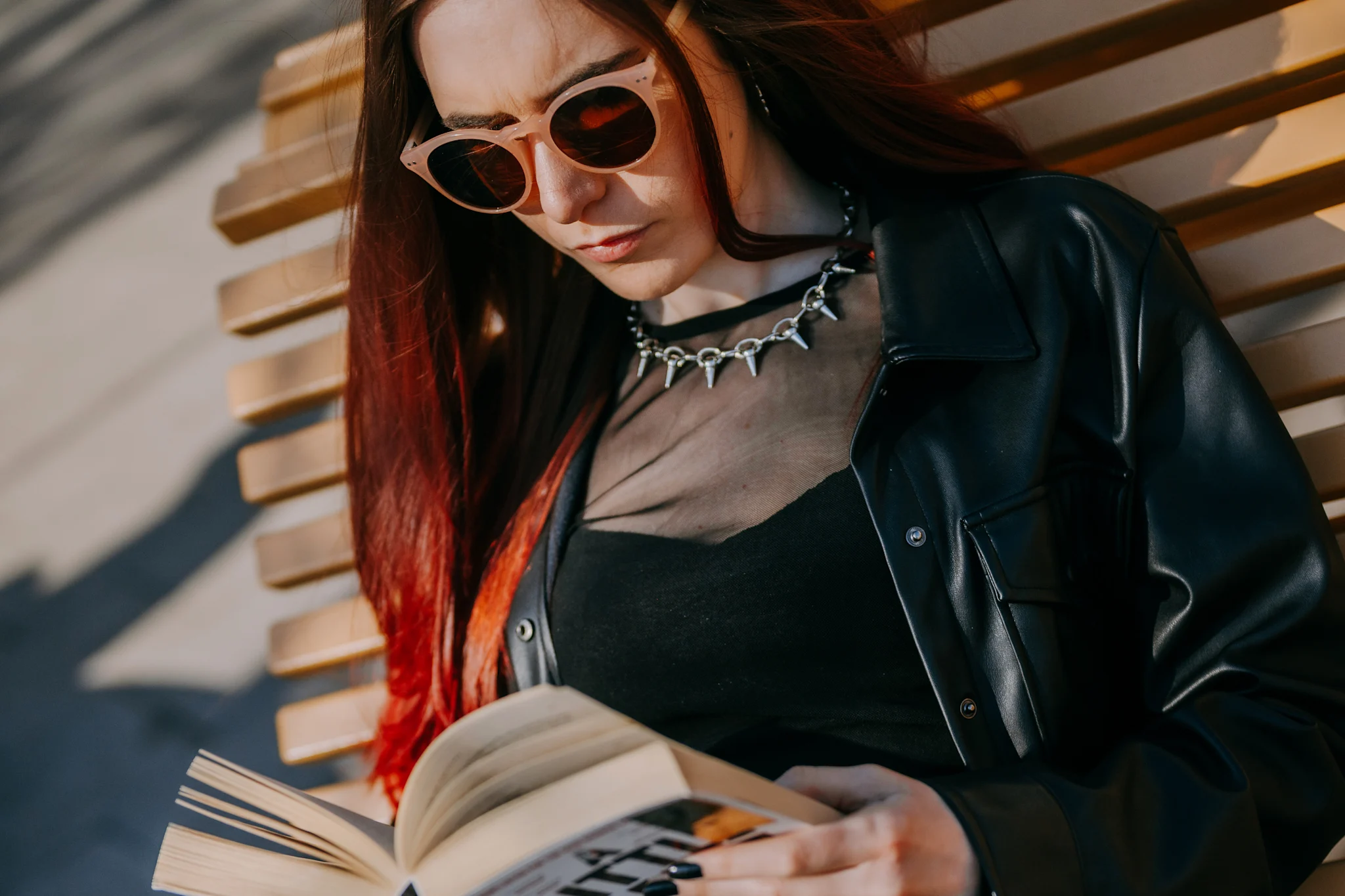 En person sitter på en benk og leser en bok, iført en svart jakke og med langt rødt hår.
