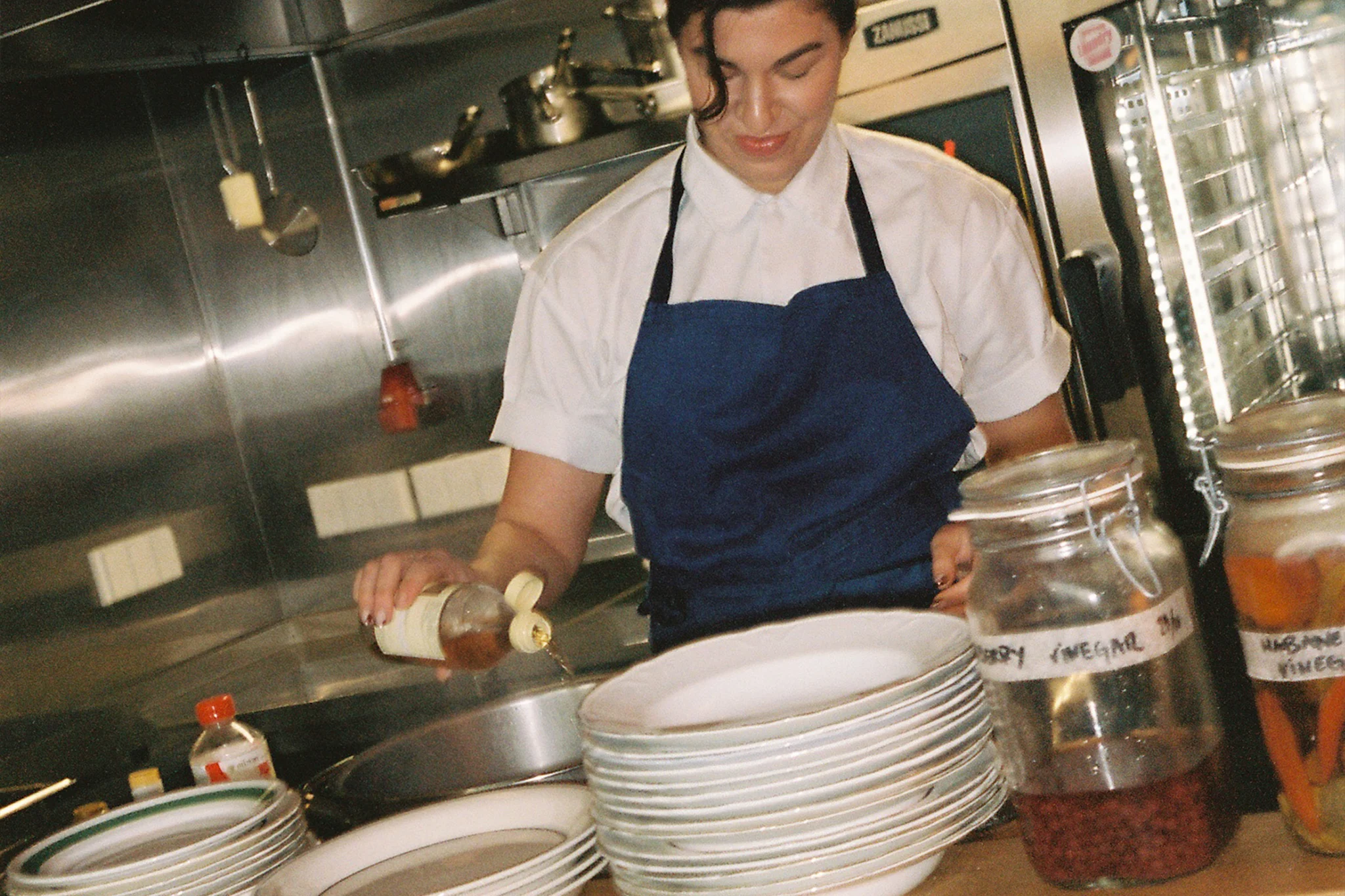 Henkilö keittiössä kaataa etikkaa kulhoon, taustalla pino lautasia.