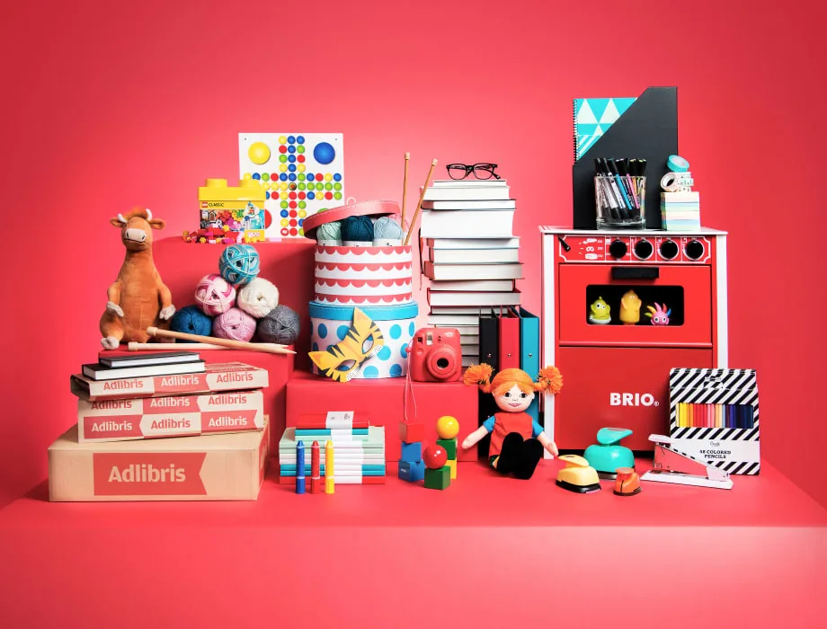 Kokoelma leluja ja askartelutarvikkeita punaisella taustalla, mukaan lukien kirjoja, rakennuspalikoita ja leikkikeittiö.