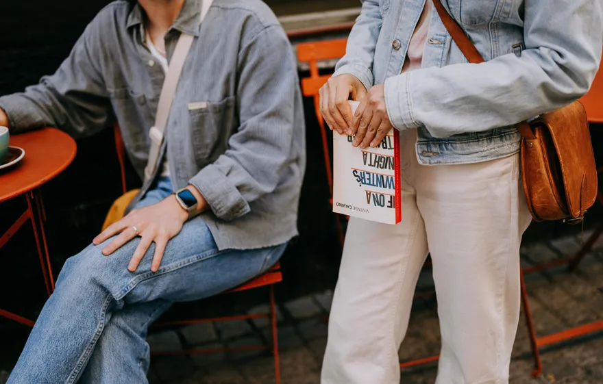  Två personer i jeanskläder sitter och står vid ett orange cafébord utomhus; en av dem håller en bok i handen.