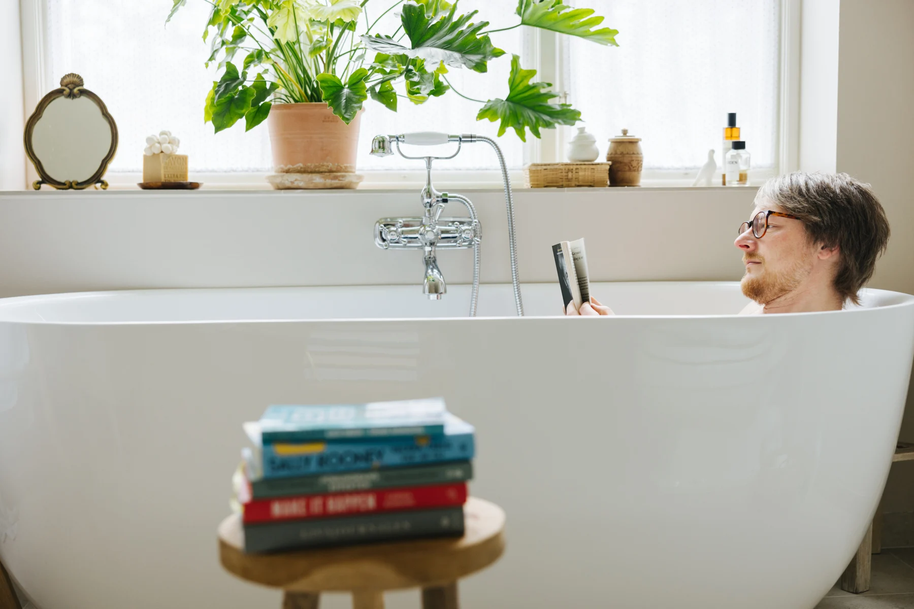 Person som leser en bok i et badekar med planter i bakgrunnen.