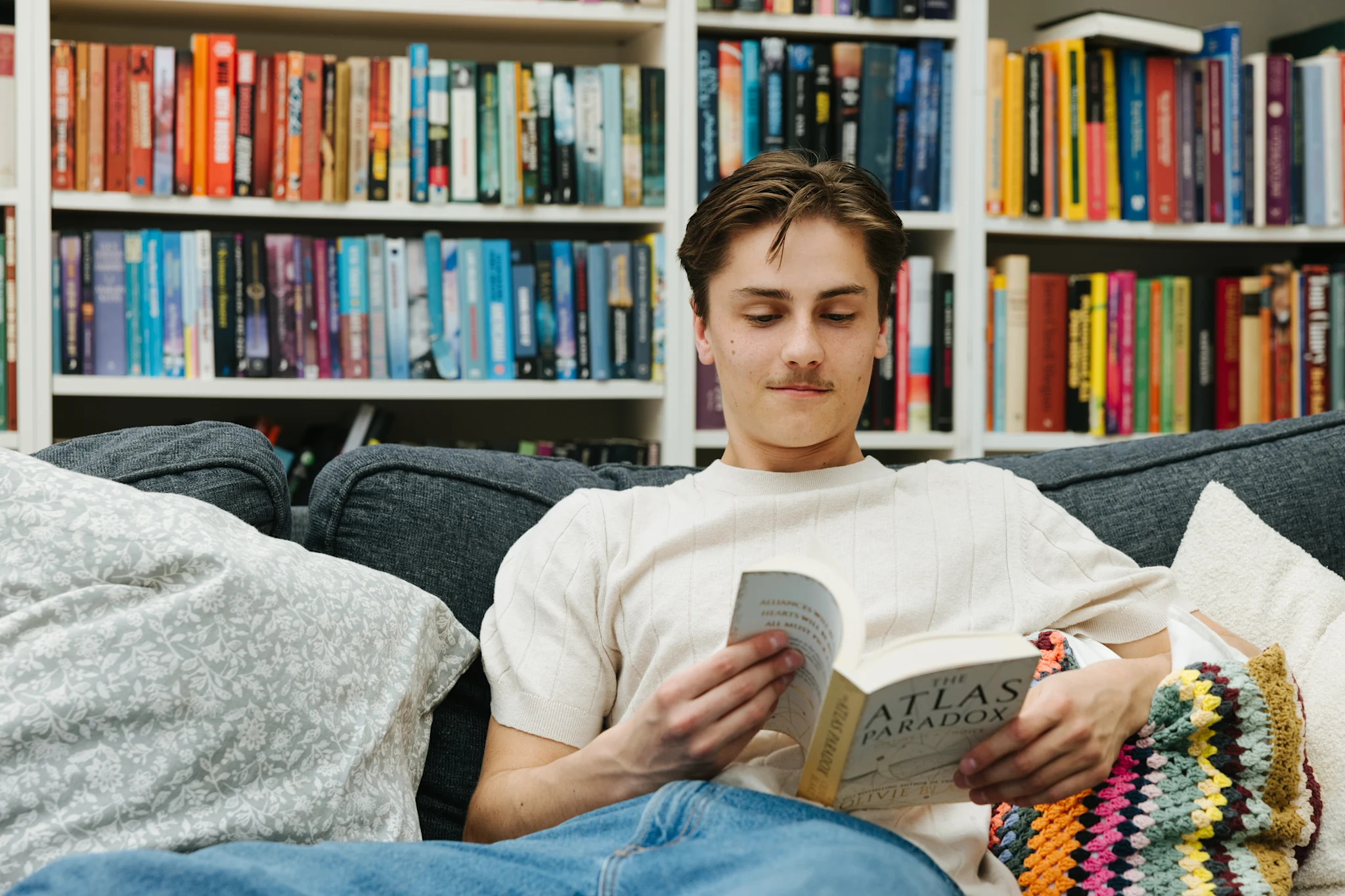 En person som sitter på en soffa och läser en bok, omgiven av en bokhylla med färgglada böcker.
