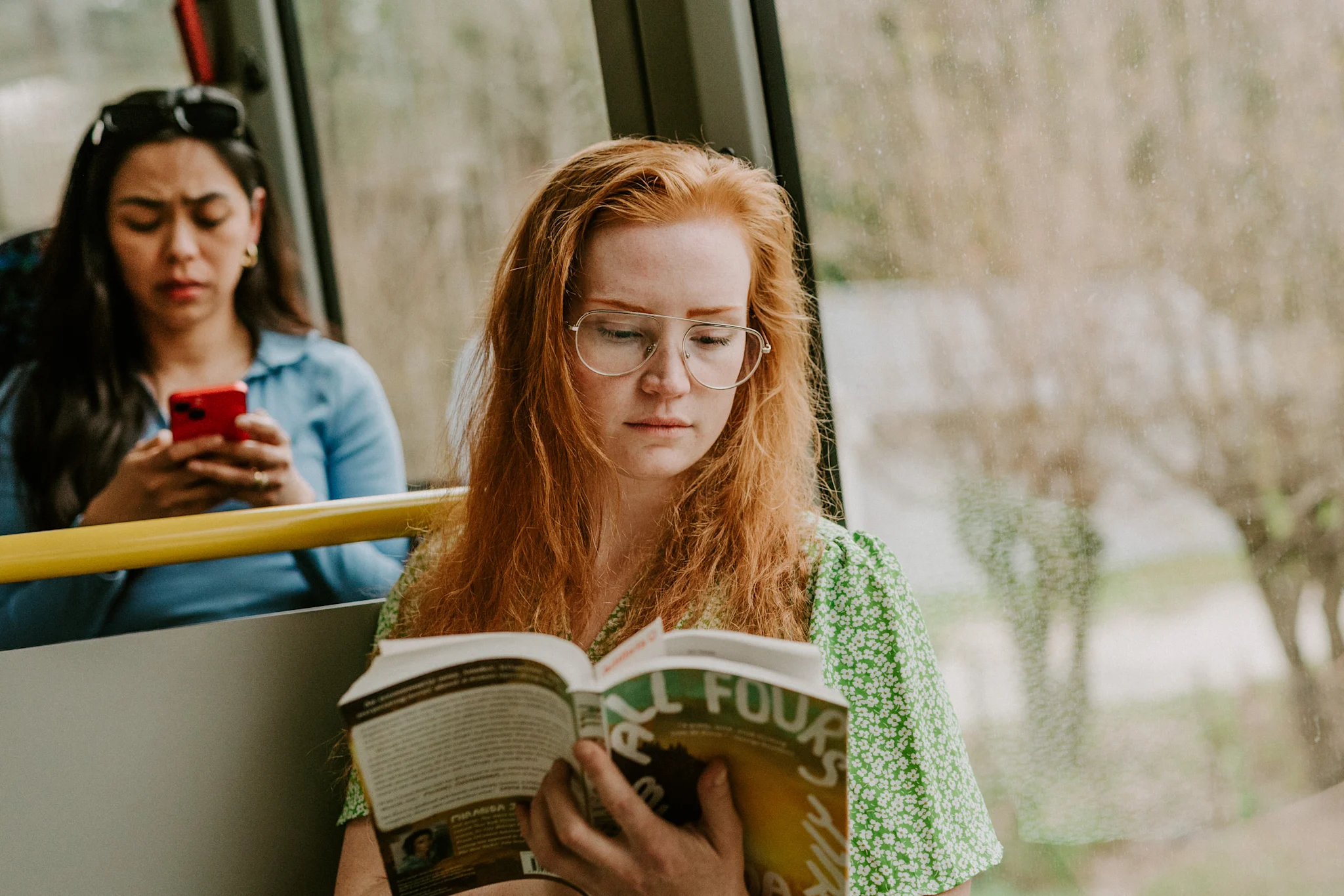 En person sitter i en buss og leser en bok iført en grønn blomstret topp.