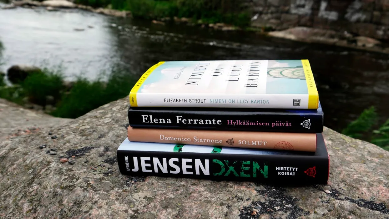 Neljä kirjaa pinoittain kivellä joen rannalla, ympärillä vihreyttä.