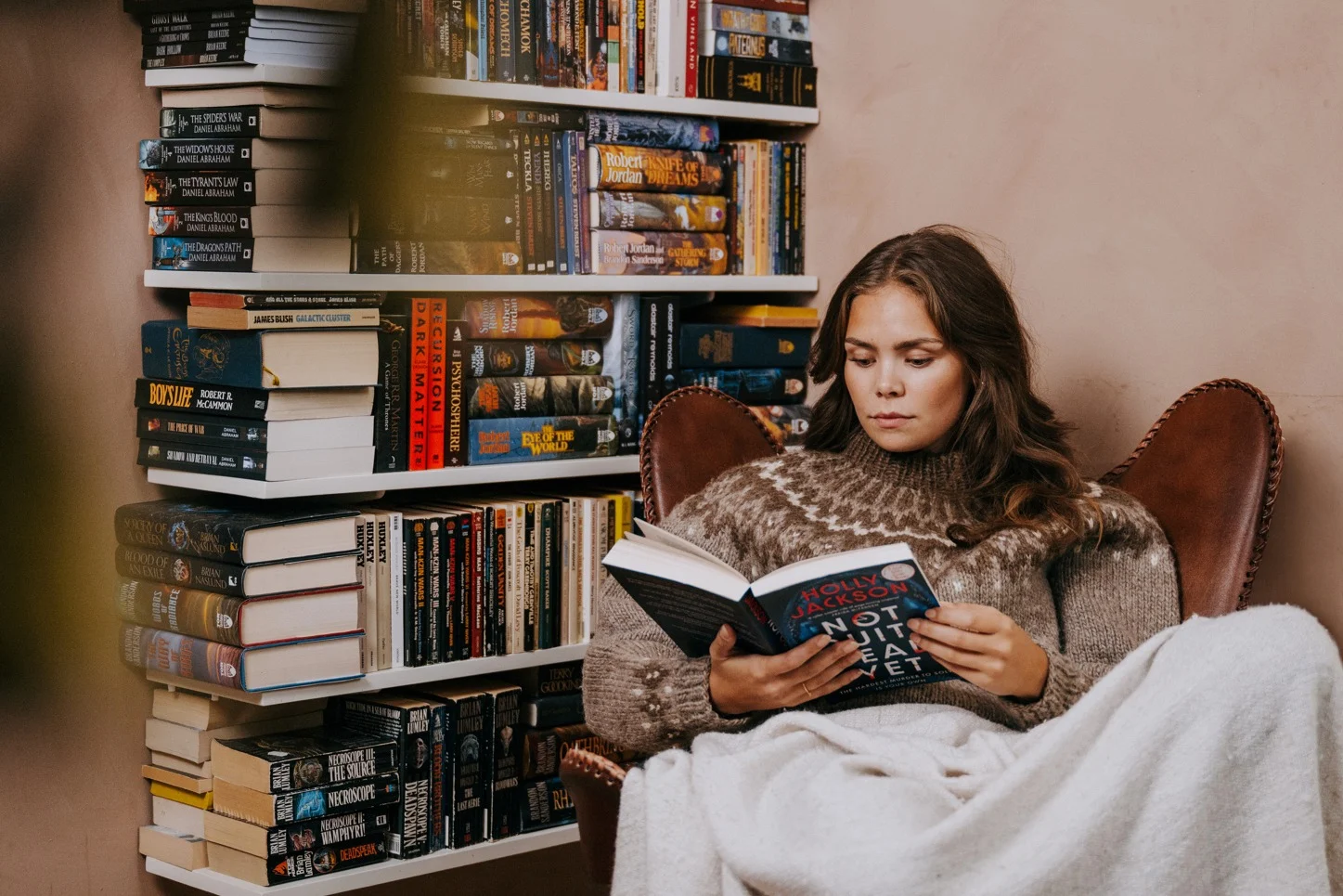 Kvinna sitter i en fåtölj med en filt och läser en bok, omgiven av bokhyllor fyllda med böcker.