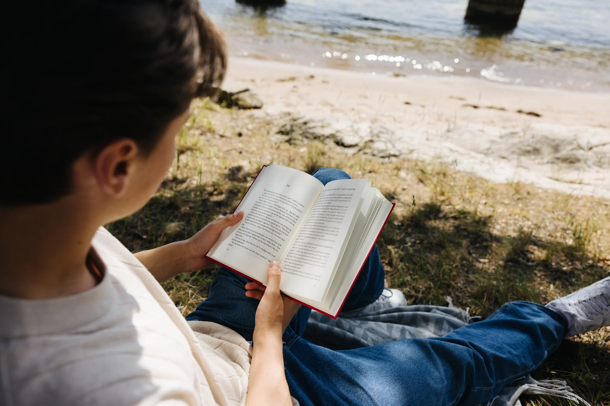 En person läser en bok vid vattnet, omgiven av grönska och sandstrand.