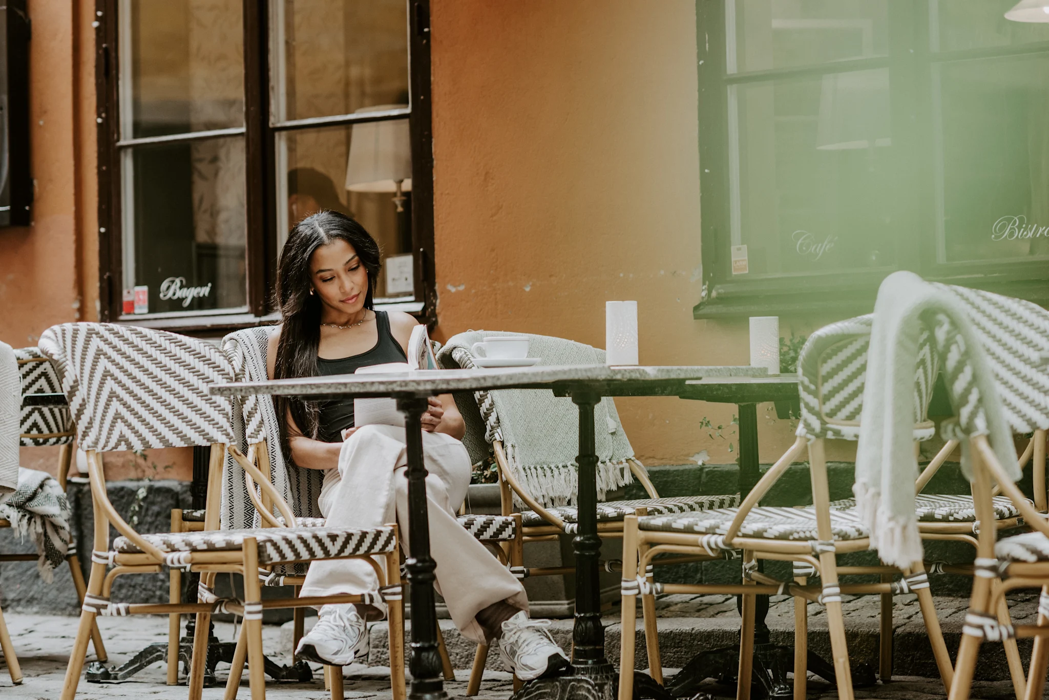 En person sitter vid ett cafébord och läser i en bok utomhus på en gata med stolar och en vägg i bakgrunden.