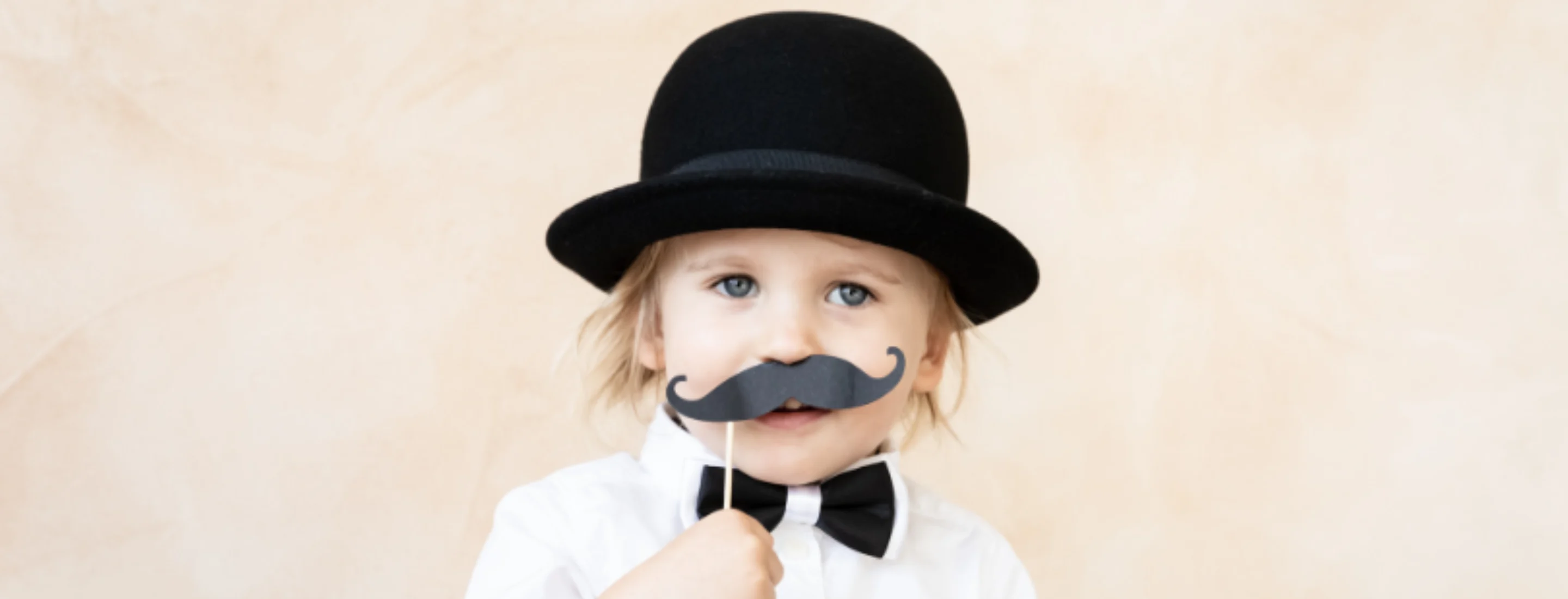 A child wearing a bowler hat and holding up a fake moustache on a stick to their upper lip