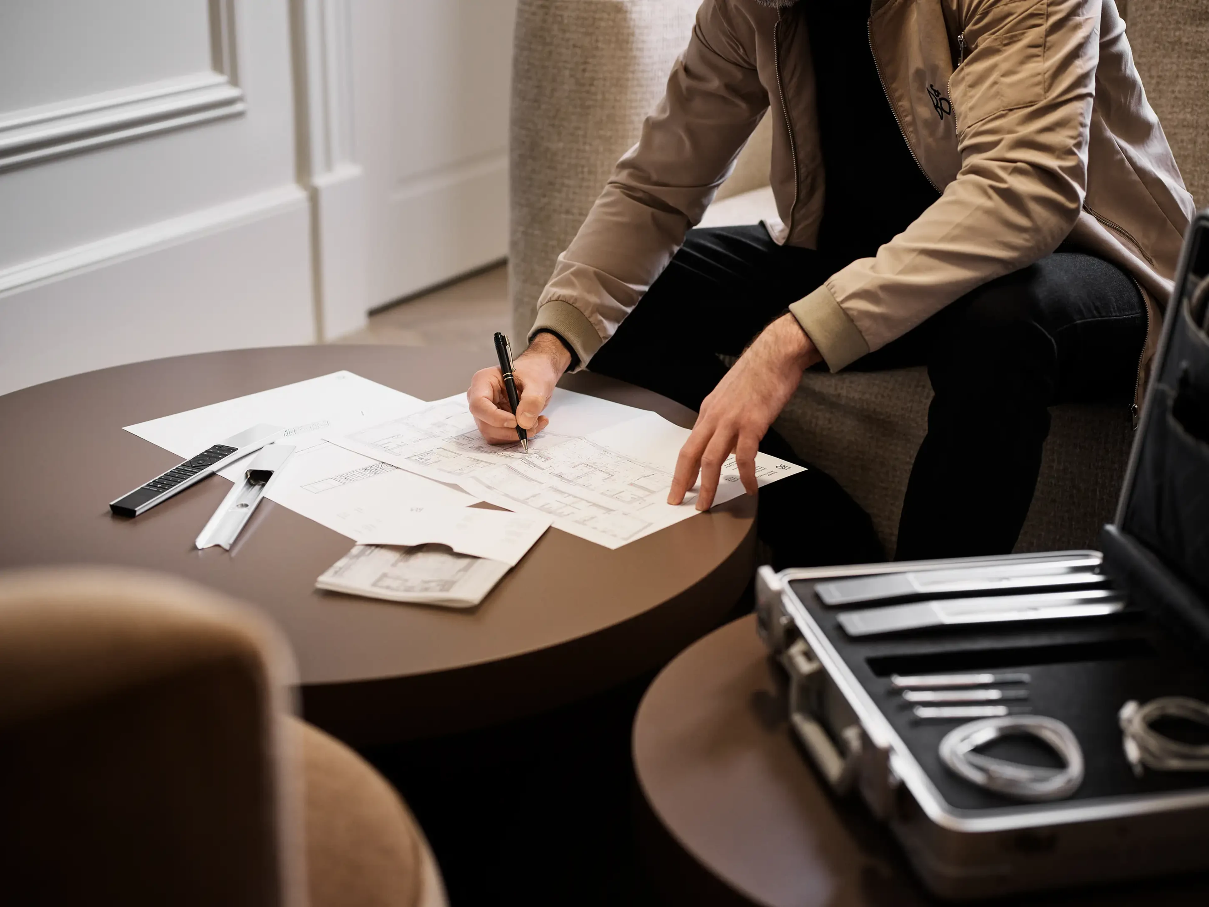 Interior designer marking up floor plans on a coffee table, with a Bang & Olufsen remote and product samples nearby