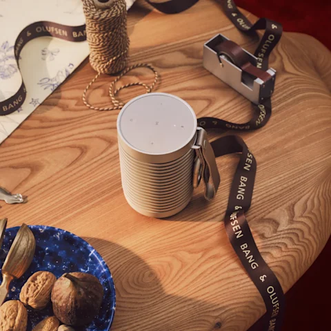 Bang & Olufsen Beosound Explore speaker on wooden table with branded ribbon, twine spool, and blue plate with nuts.