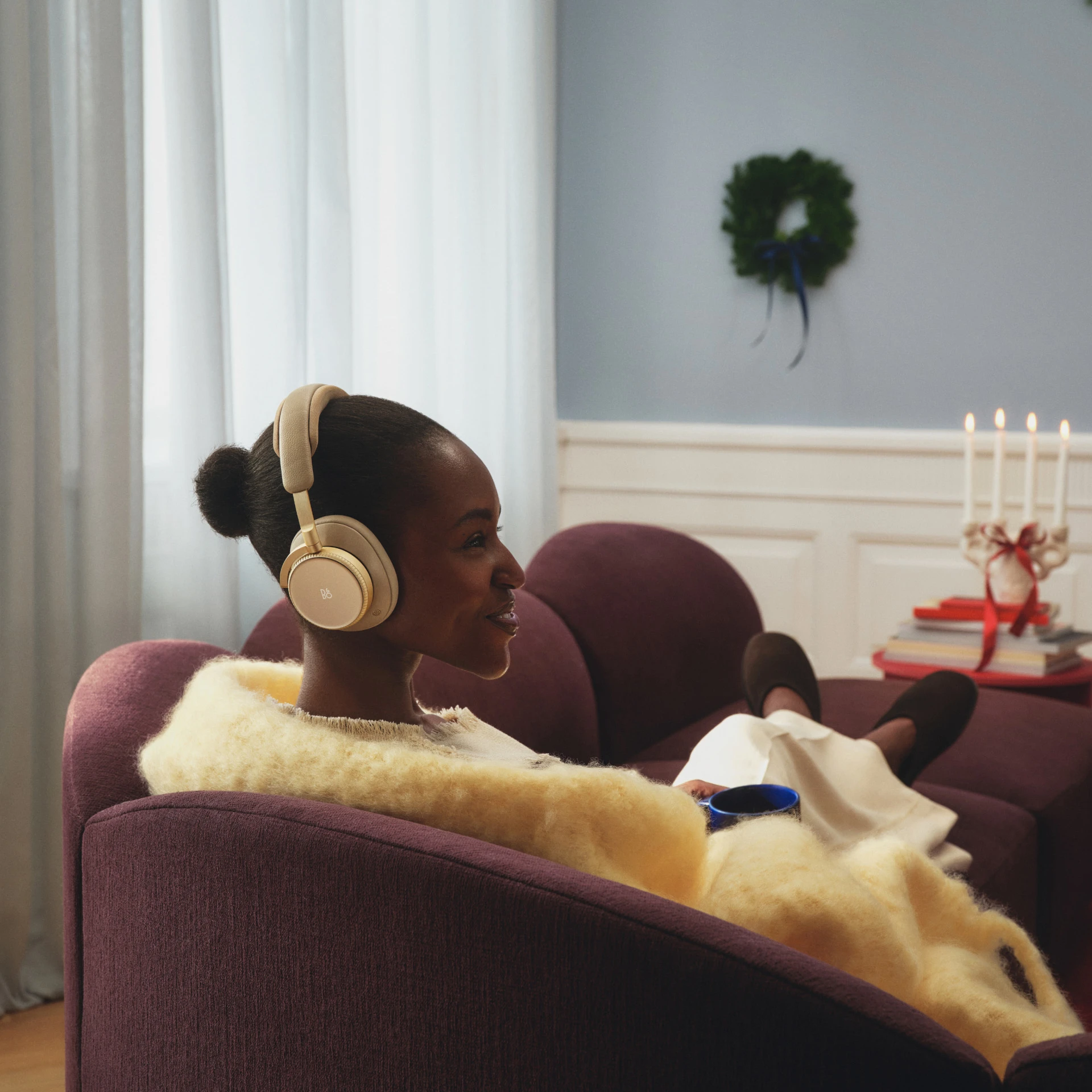 Person wearing gold Beoplay H100 headphones relaxing on purple couch with blanket in cozy room with candles and holiday decor.