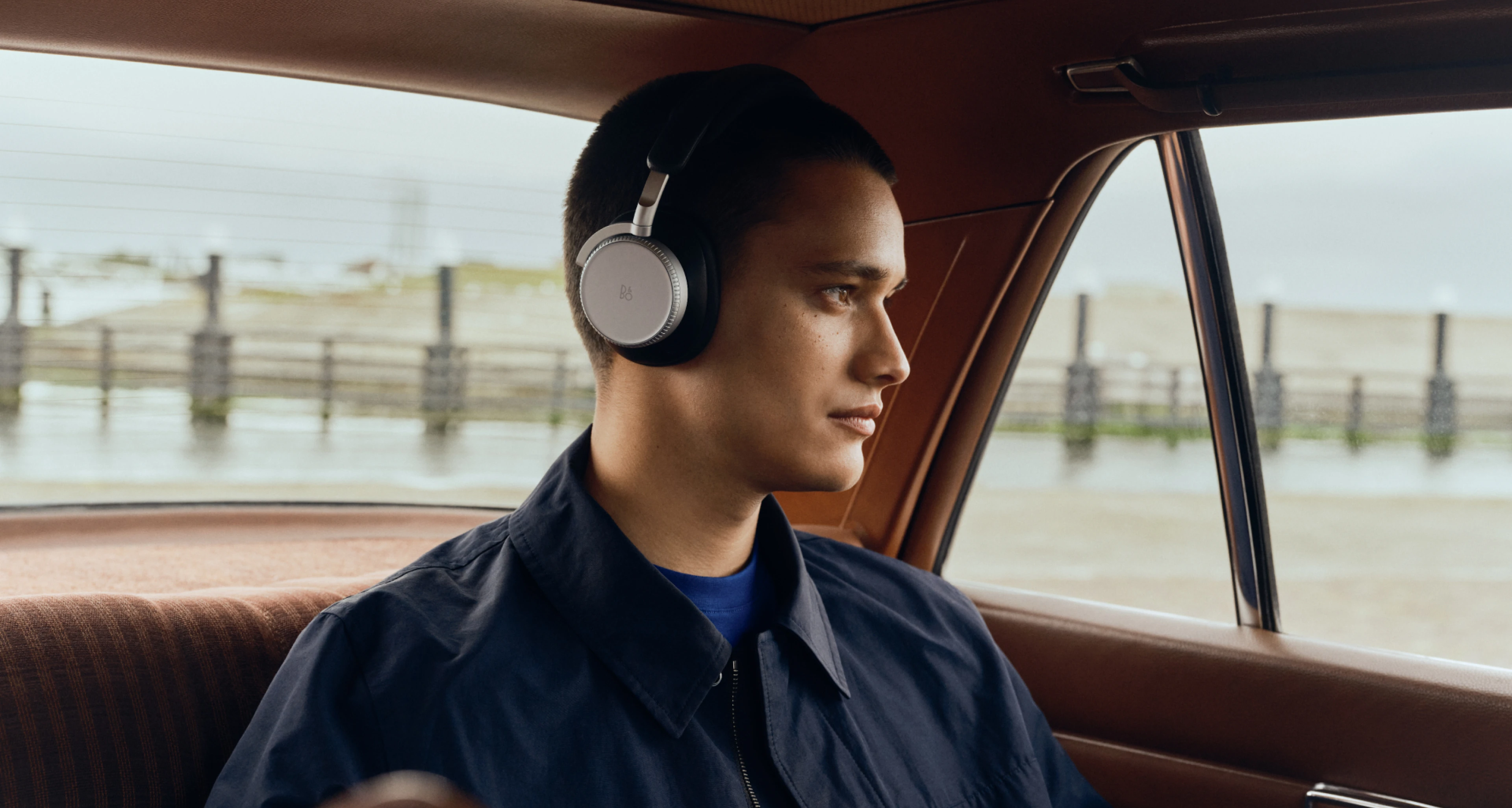 車内にいる男性の横顔。Infinite BlackカラーのBeoplay H100を装着し、窓の外を見つめながら物思いにふけっている。
