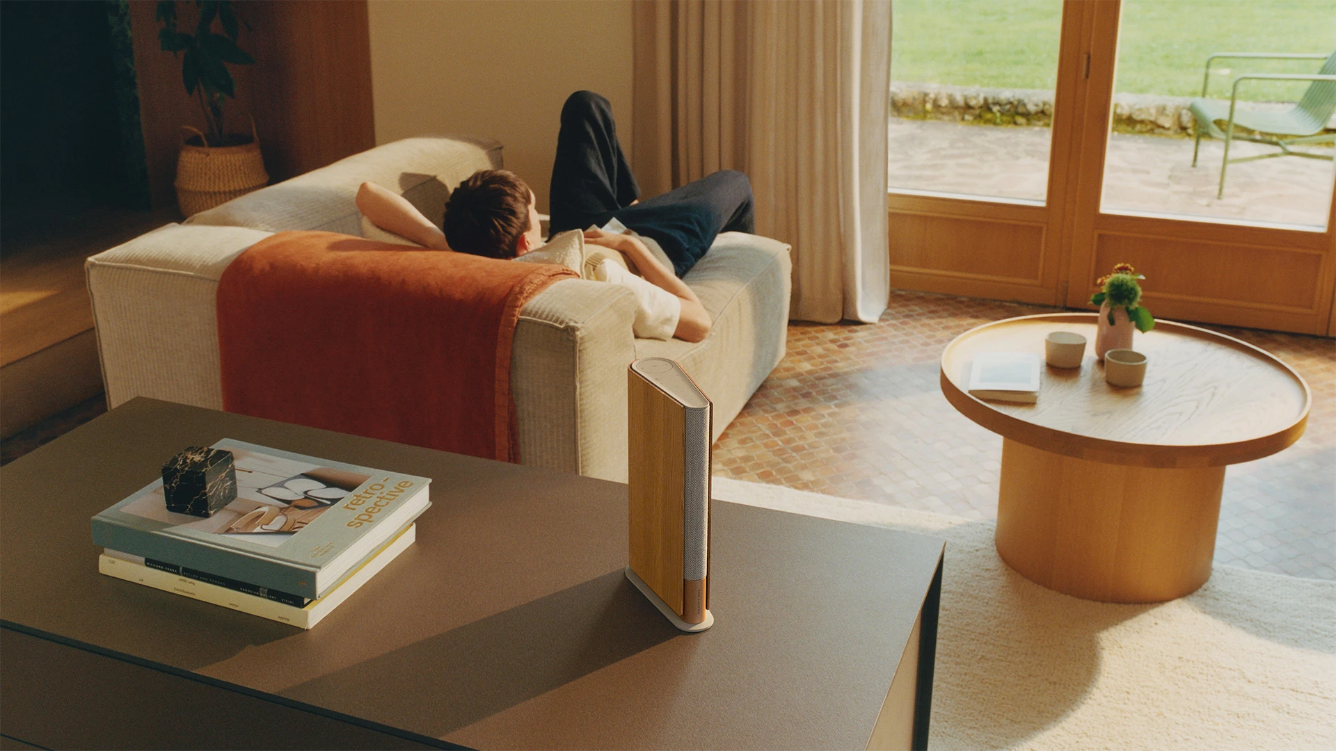 Beosound Emerge speaker on the table in a living room, and there is a man enjoying the music lying down on the sofa