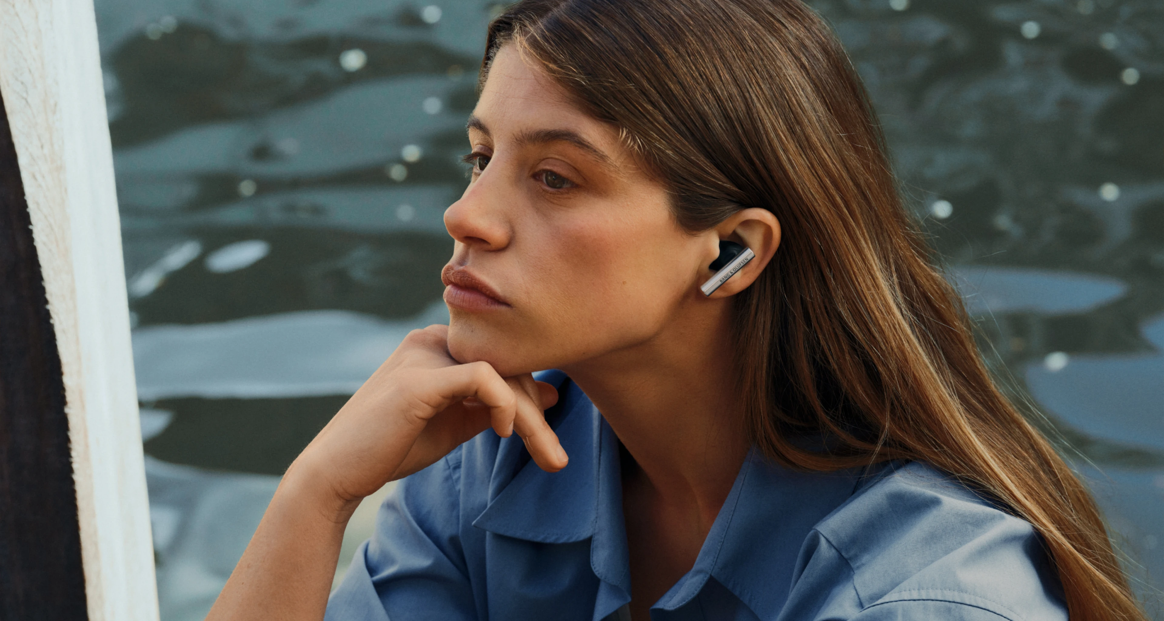 AluminiumカラーのBeo Graceイヤーバッドをつけ、水辺に静かに座っている女性。視線は遠くを見つめ、音楽に包まれながら思いを巡らせている。