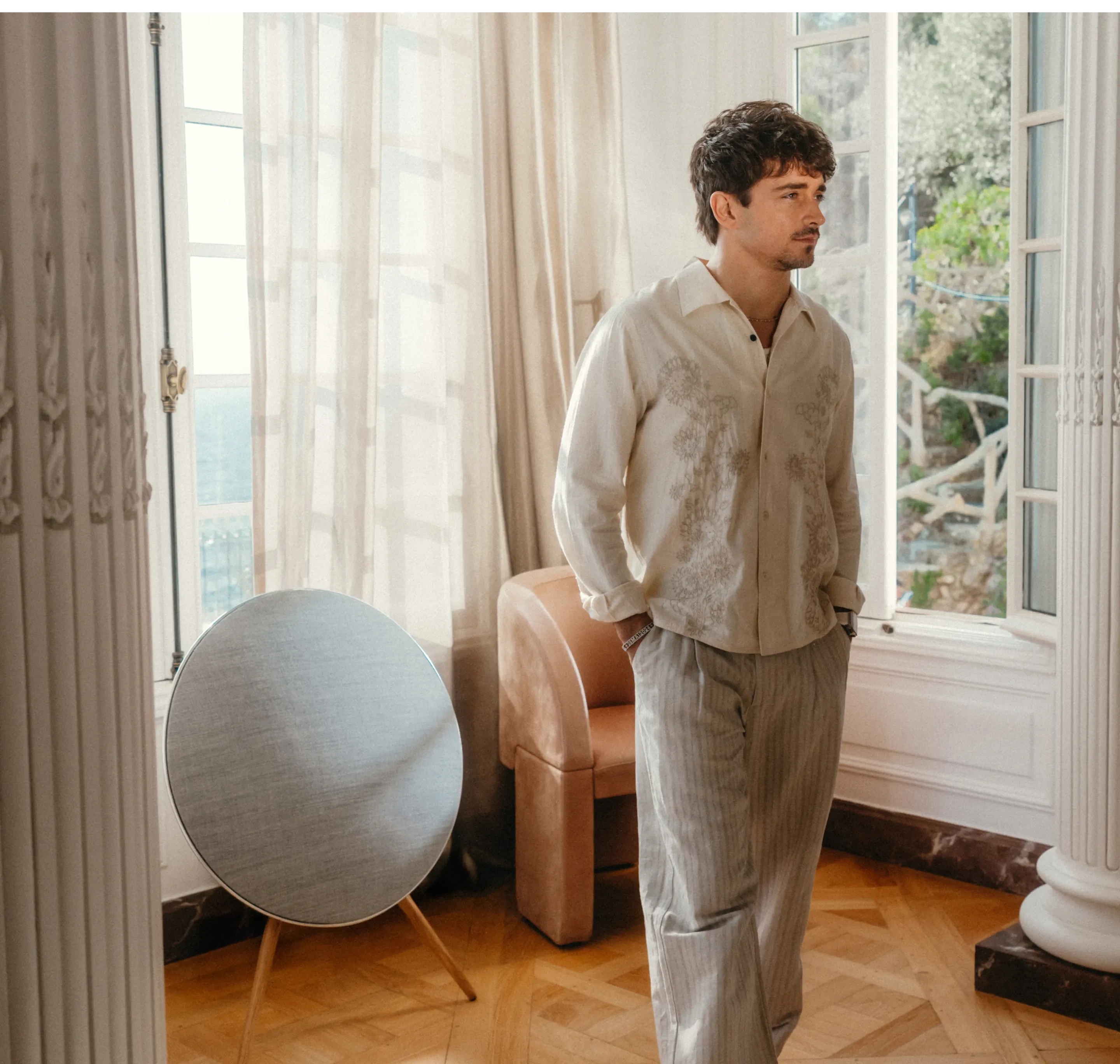Person in cream embroidered shirt and light pants standing by bright windows in elegant room with Beosound A9