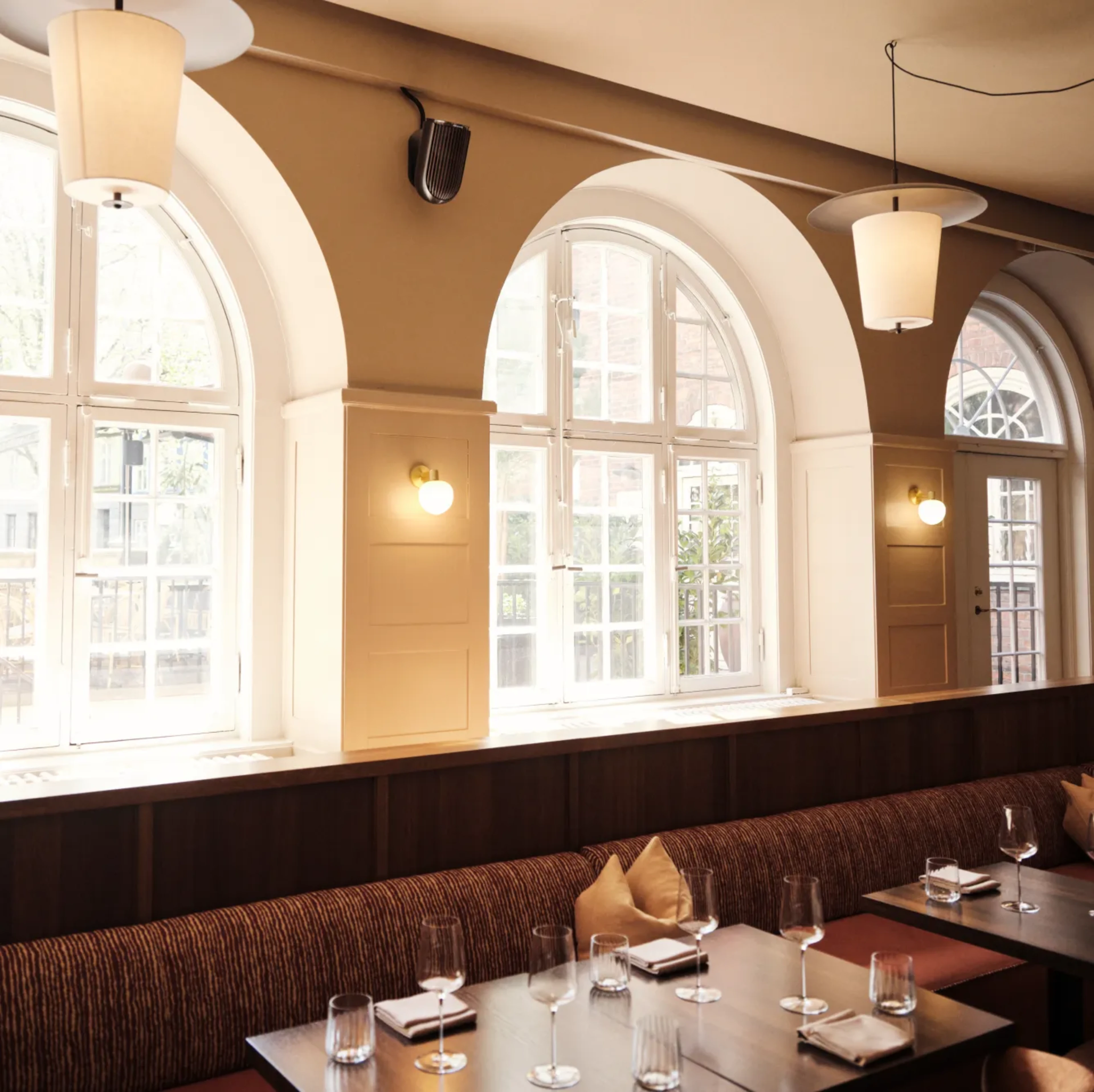 Elegant restaurant interior with arched windows, upholstered banquette seating, dark wood tables, and sculptural pendant lighting.