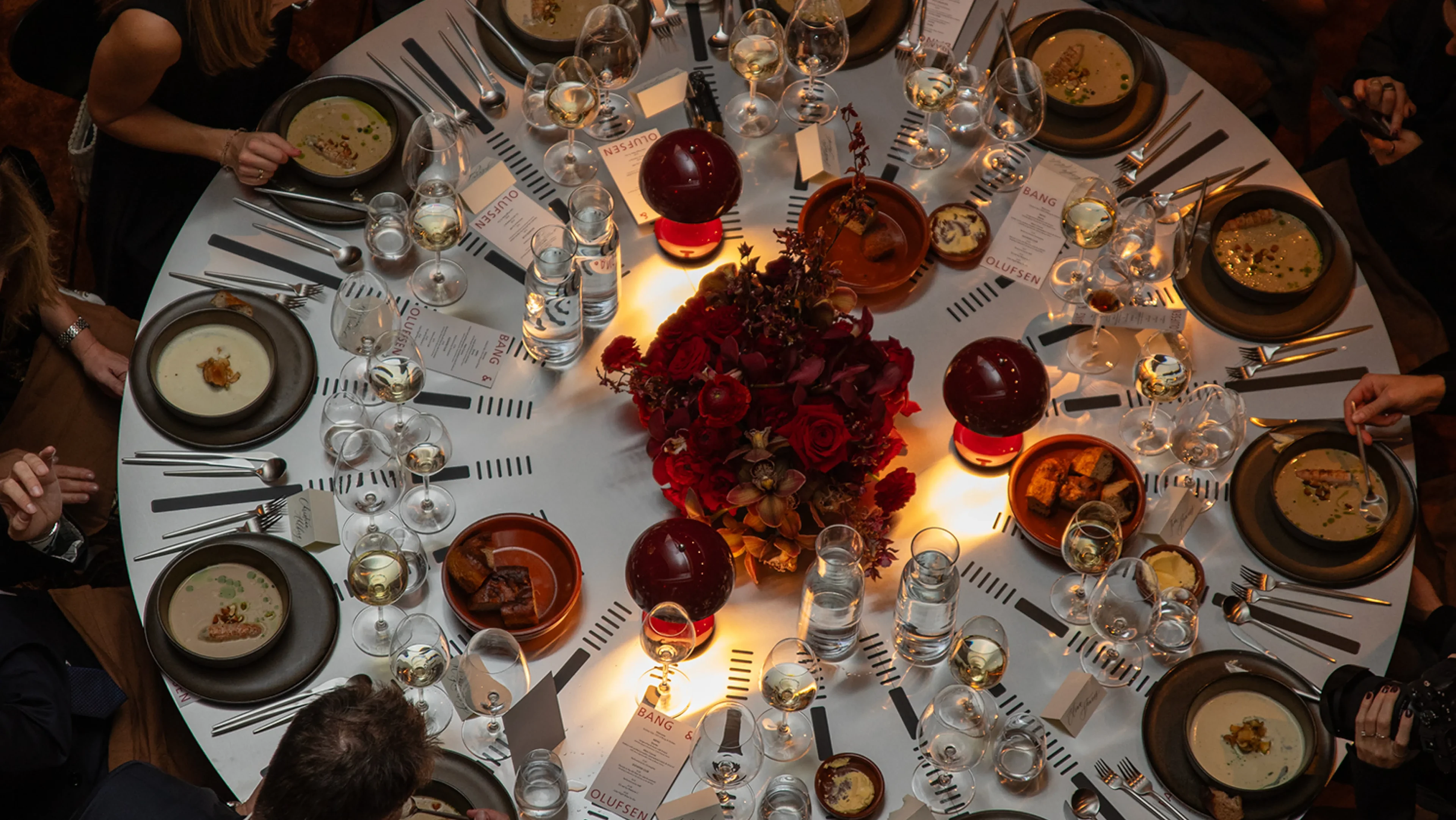 Elegant round dinner table with red floral centerpiece, wine glasses, and guests seated around enjoying a formal meal.