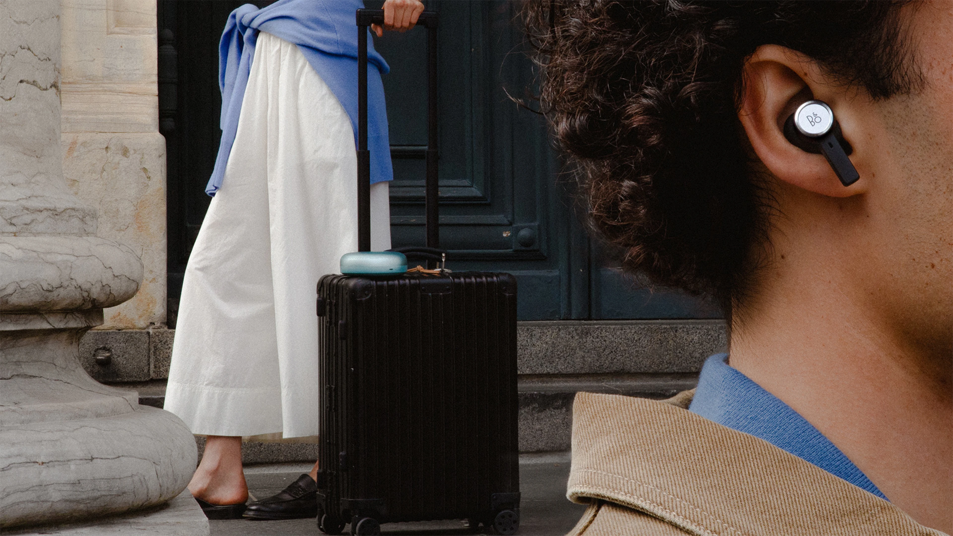 Man wearing Beoplay Eleven in the foreground and woman carrying luggage with Beosound A1 in the background.