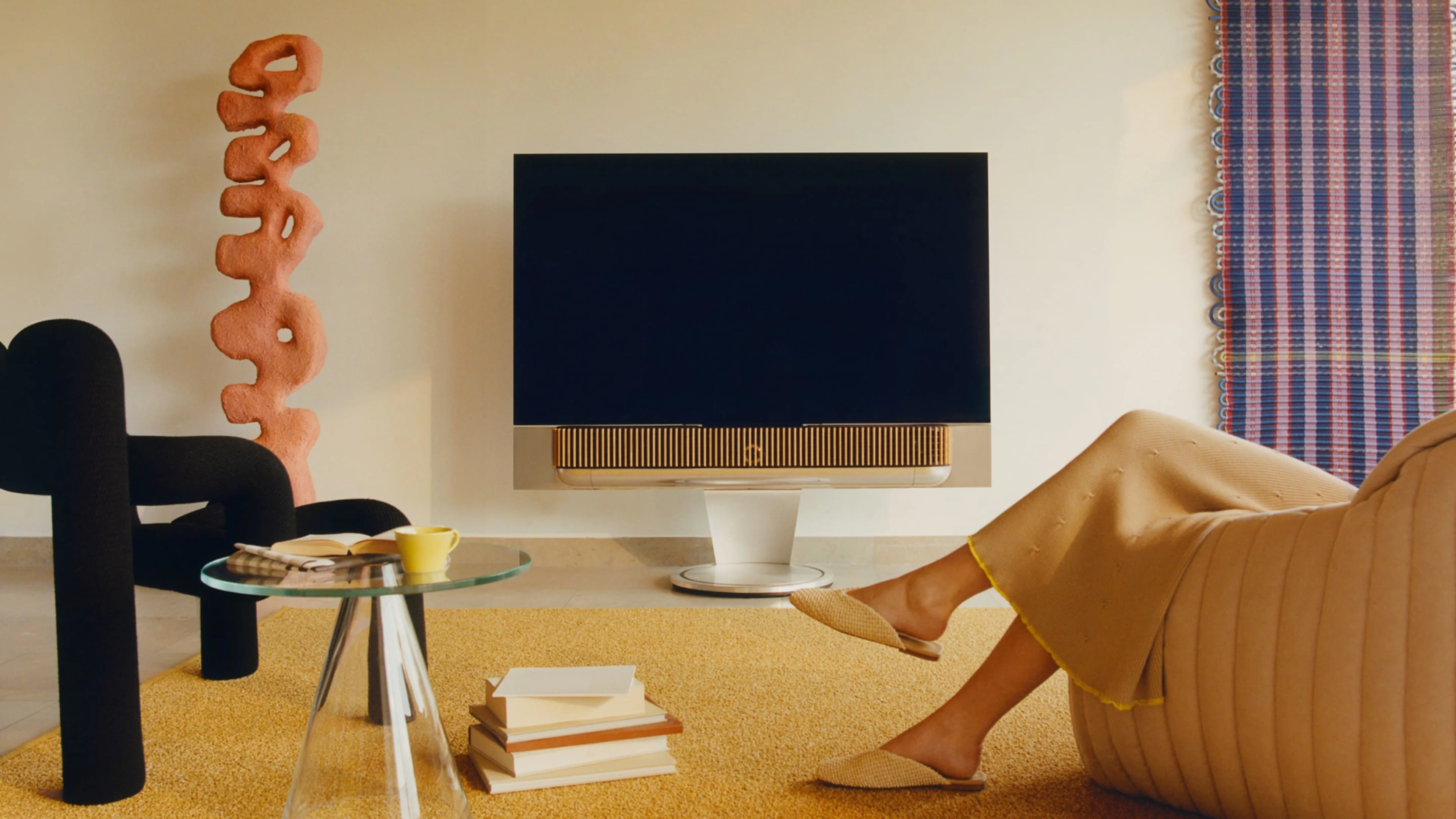 Gold tone aluminum Beovision Theatre is placed in a living room where a lady is seating on a cozy looking sofa