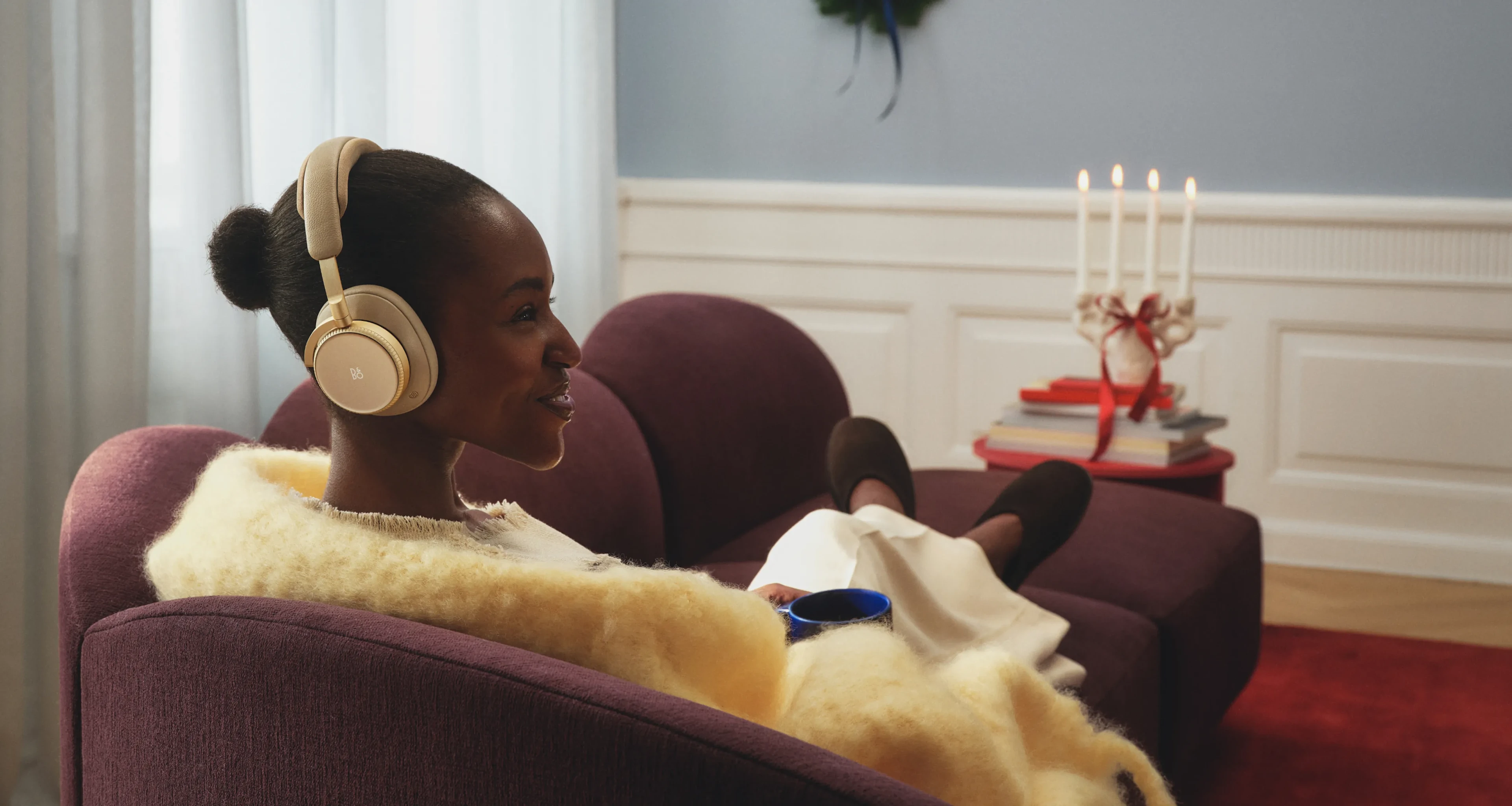 一位女子配戴 Beoplay H100 Hourglass Sand 坐在沙發上，一邊品茶一邊聆賞音樂