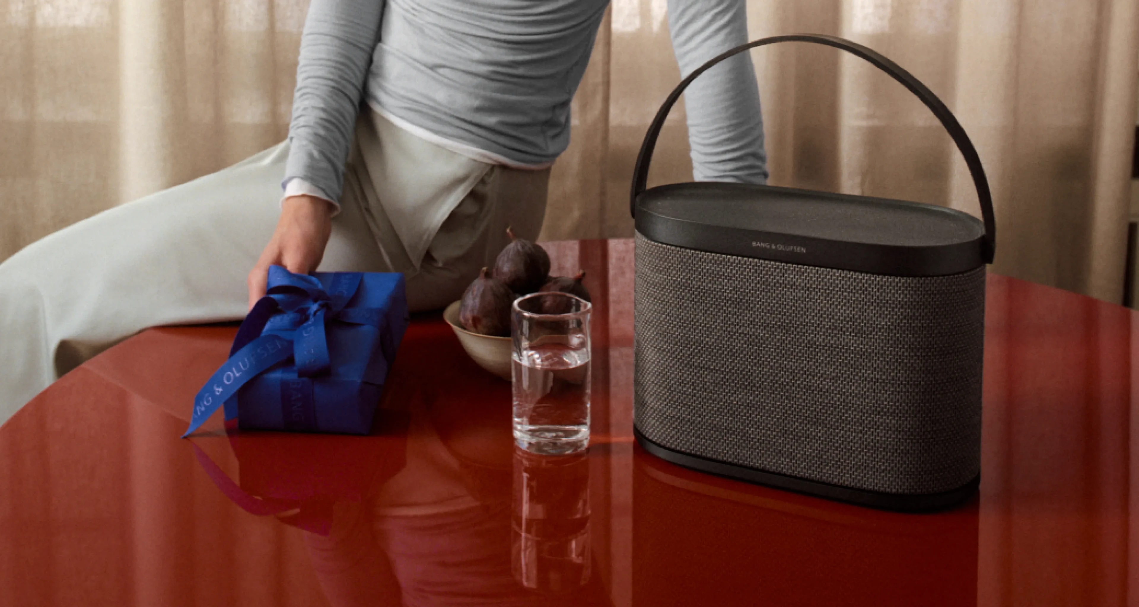Beosound A5 portable speaker in Charcoal Knit placed on a glossy red table beside a glass of water and a wrapped gift, with a person reaching across the table in a softly lit indoor setting.