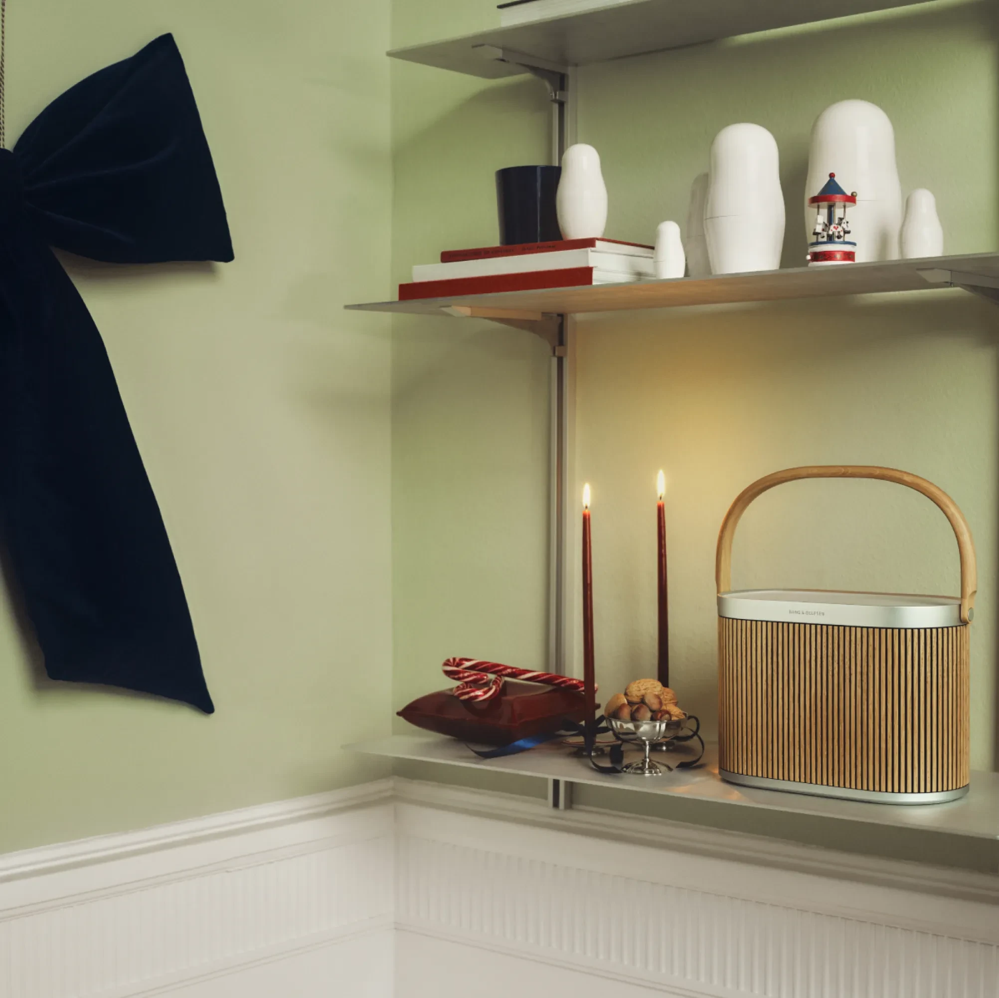 Stylish shelf display with white nesting dolls, lit candles, Beoosund A5 oak speaker, and decorative items against pale green wall.