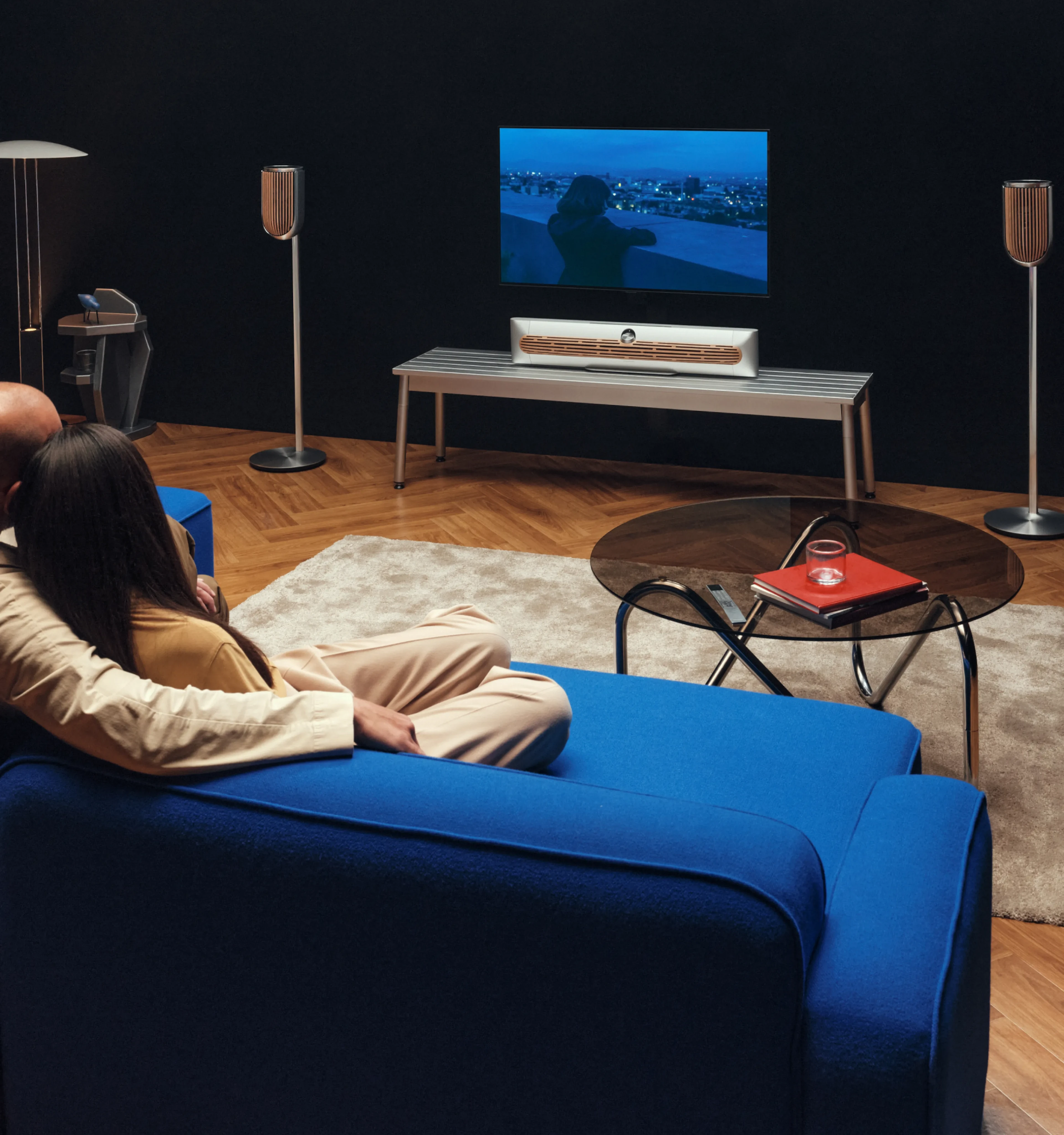 一对夫妇坐在沙发上,用他们的 Beosound Premier 扬声器和 一对 Beolab 8 看电视,我们从斜后方观察他们