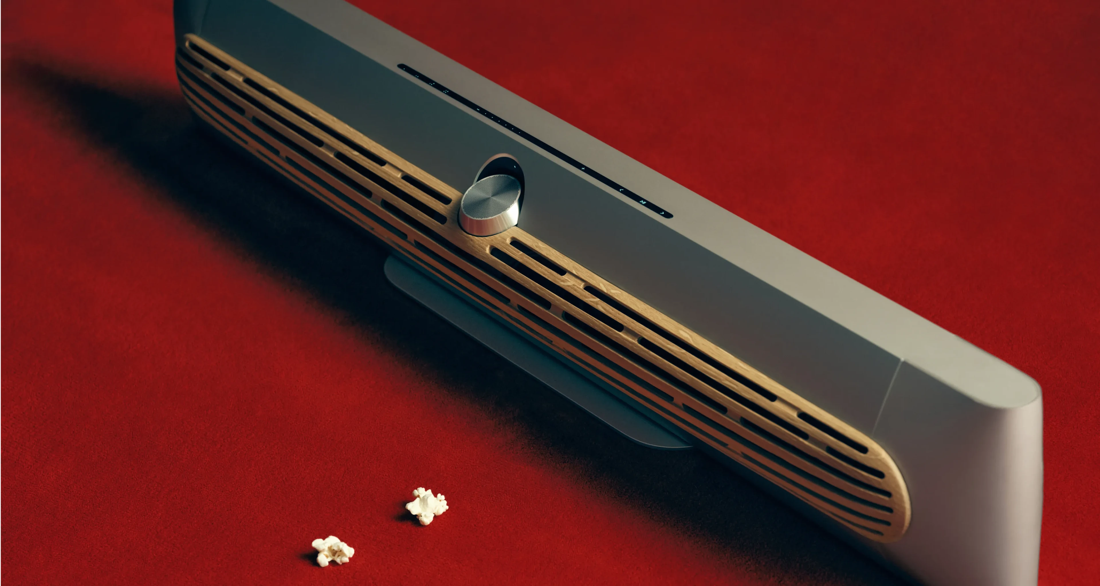 红地毯上摆放着 Beosound Premiere Natural 色款 Soundbar,配橡木外壳,前面放着两粒爆米花