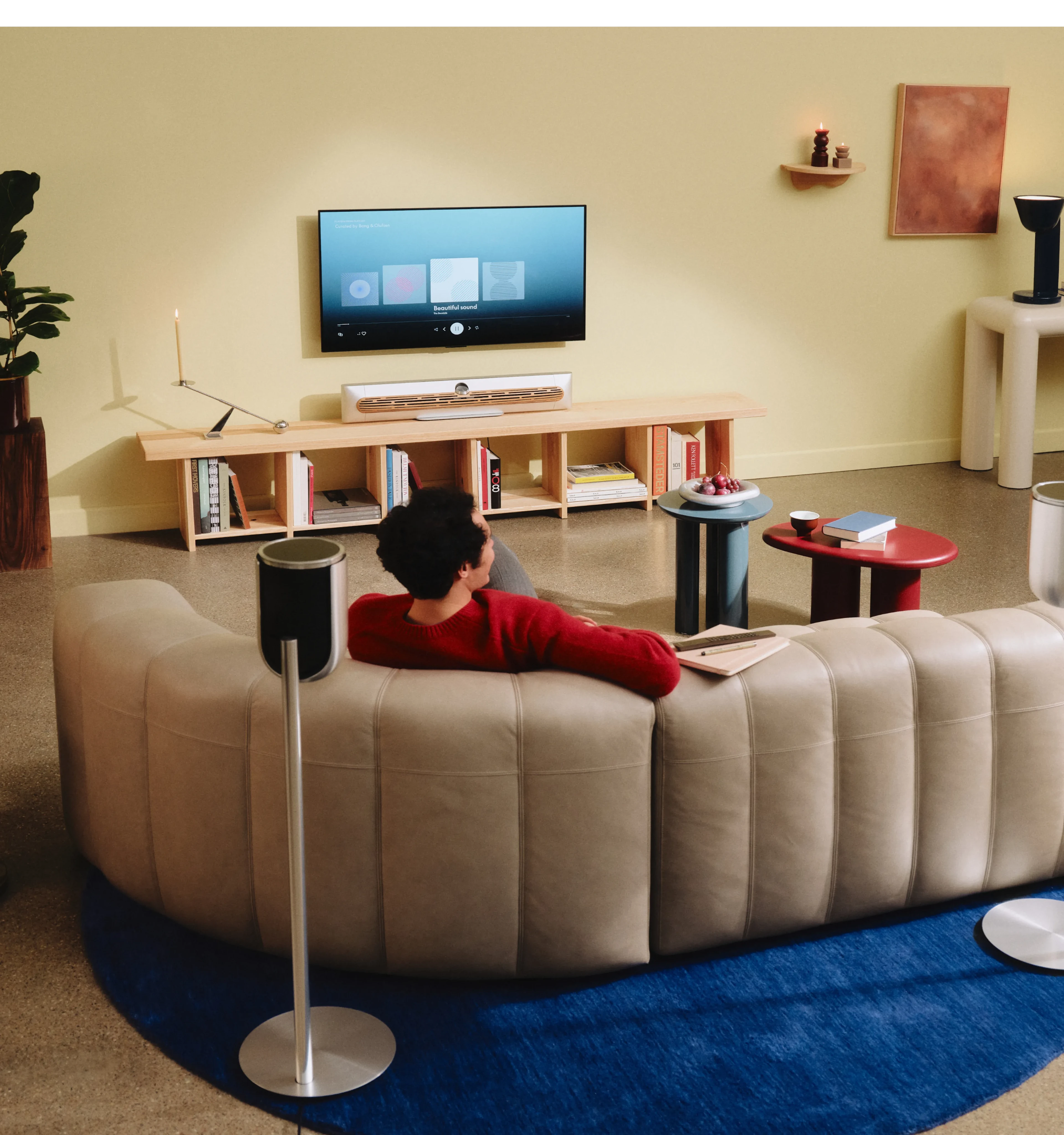 Modern living room with cream sofa, wall-mounted TV, Beosound Premiere soundbar, Beolab 8 speakers, wooden media console, and colorful accent tables on blue rug.